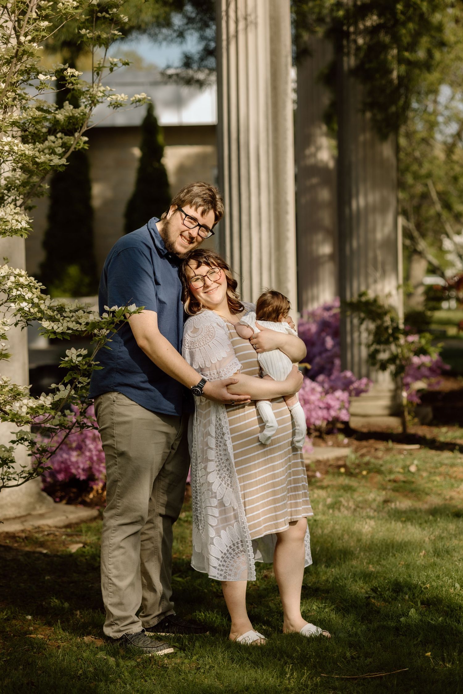 A young family poses outdoors near blooming flowers, with parents smiling as mother holds their newborn baby.