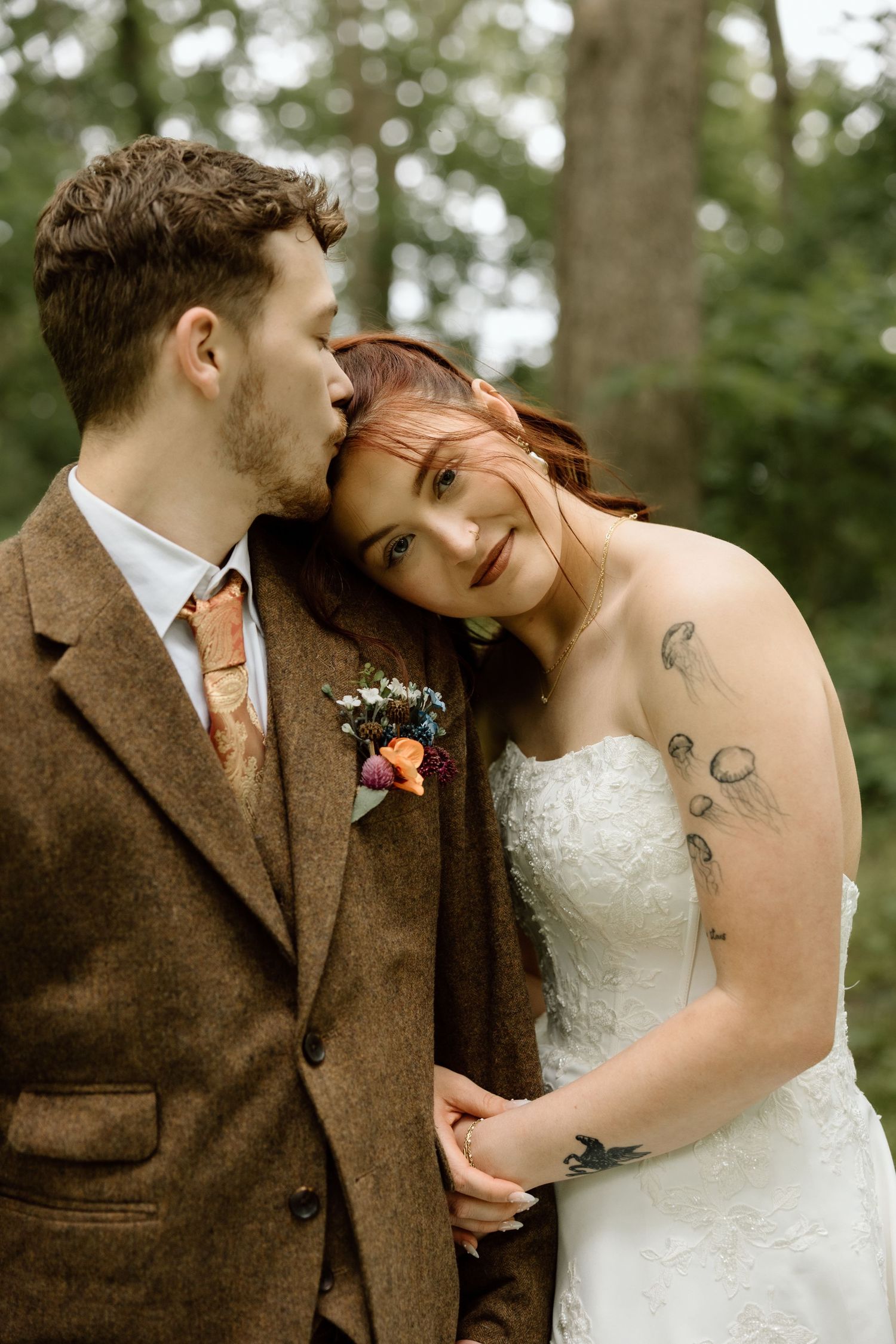 Groom in brown tweed suit kissing tattooed bride on the head during romantic outdoor wedding portraits.