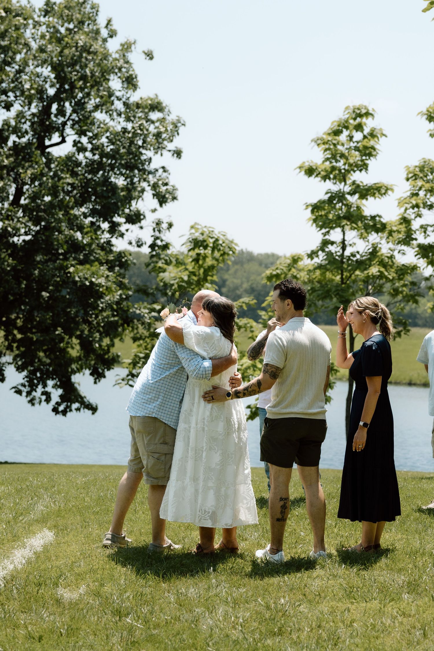 Newlyweds sharing hugs with family members on green lawn beside a peaceful lake after ceremony.
