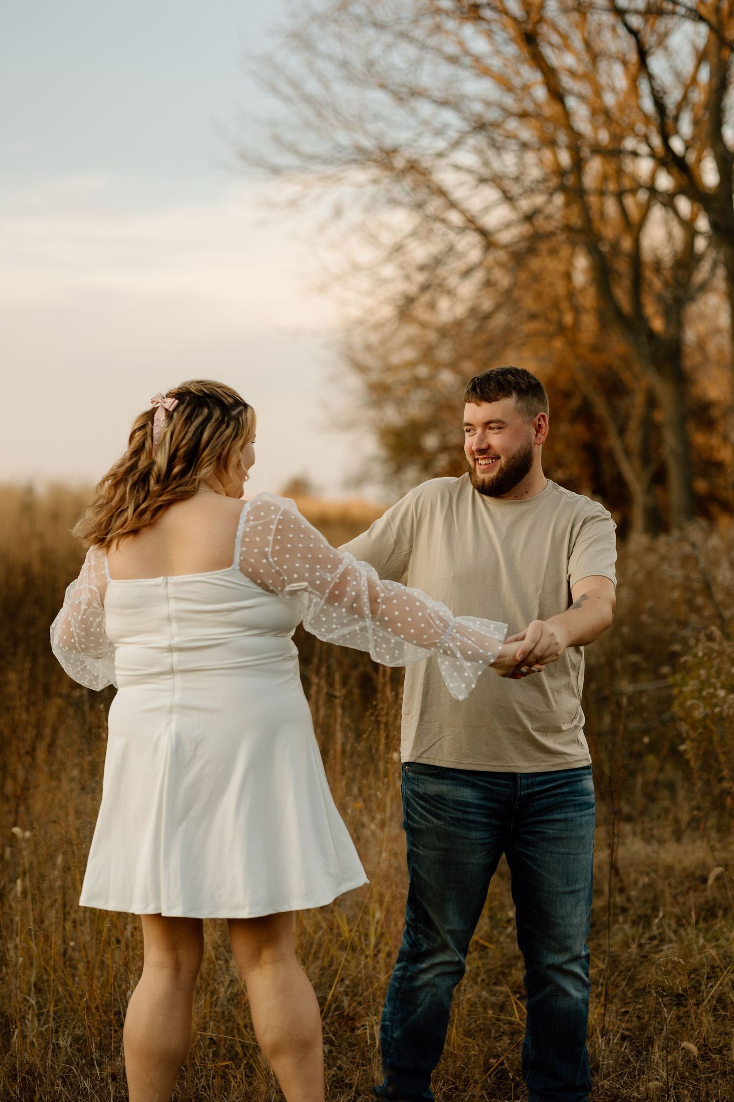Smiling bearded man in tan shirt holds hands with woman in white lace dress in autumn field at golden hour.