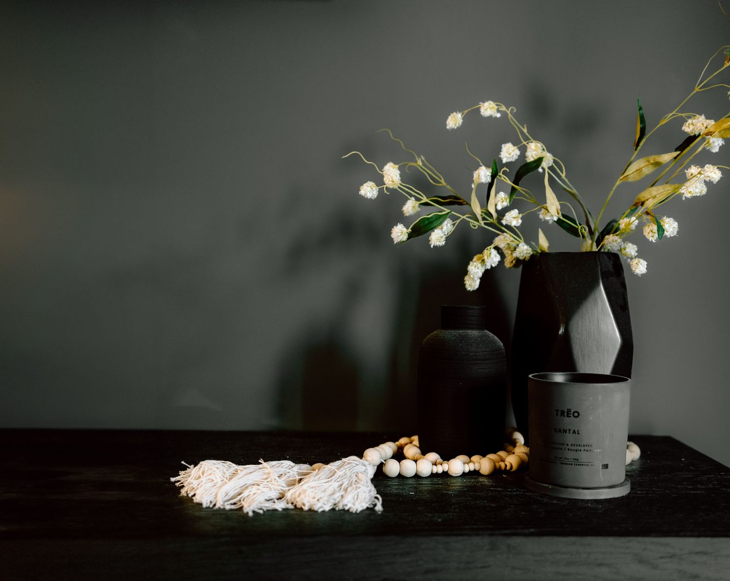 Dark moody still life with black vase, white flowers, candle, and beaded garland.
