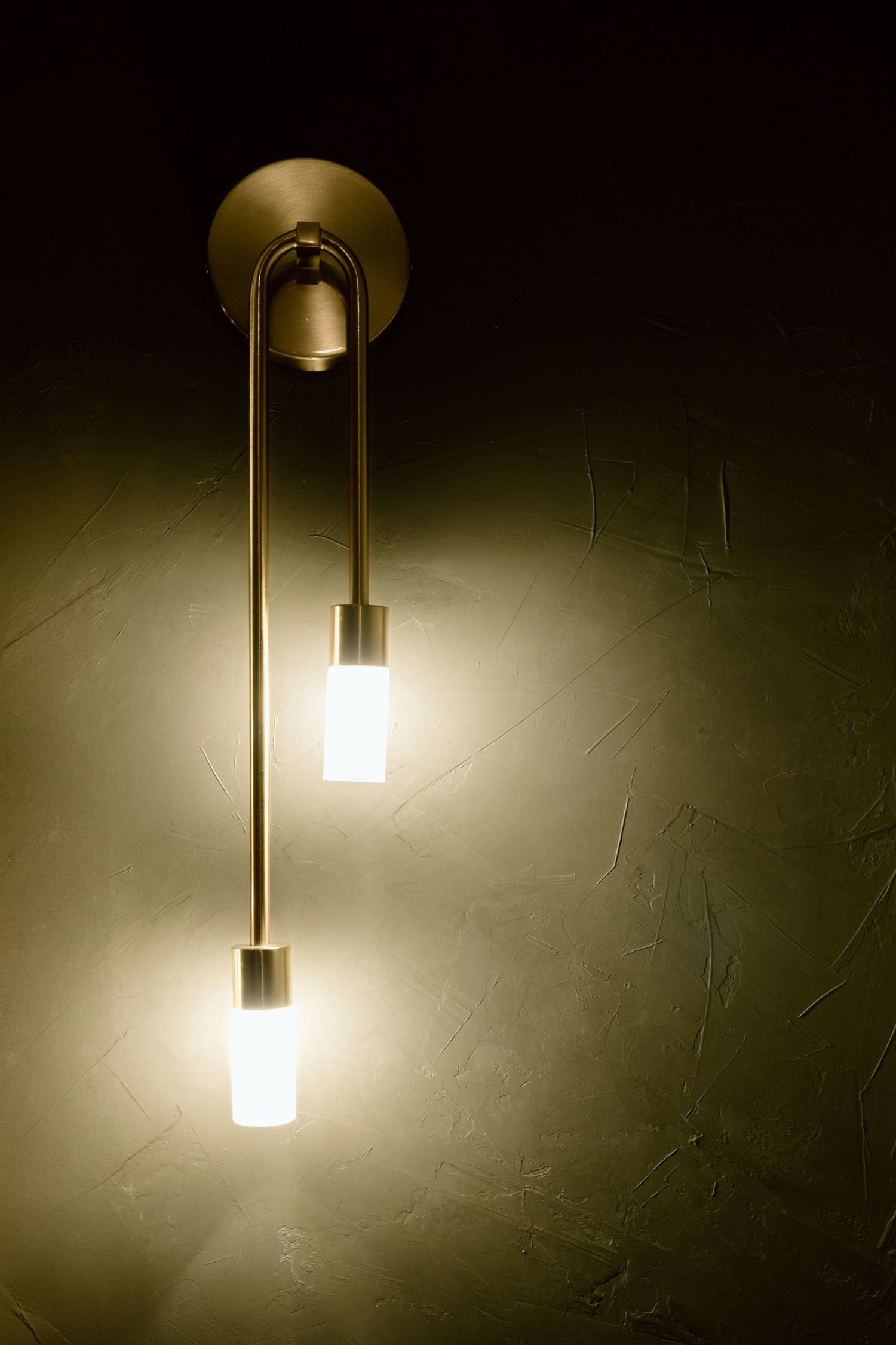 Brass double-bulb wall sconce casting warm light against a textured olive green plaster wall.