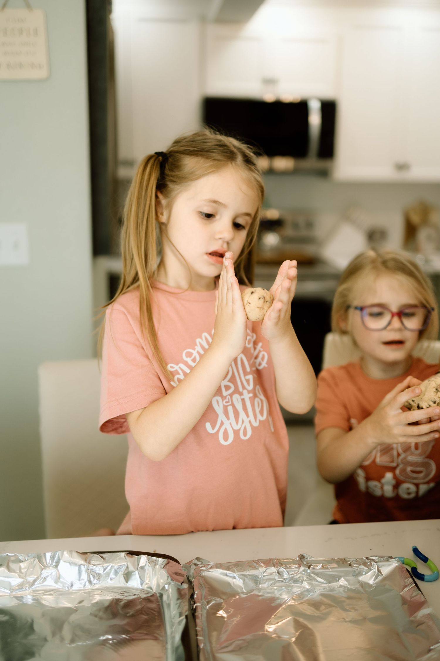 Young girl in pink shirt rolling cookie dough balls over foil-lined baking pan in a home kitchen.