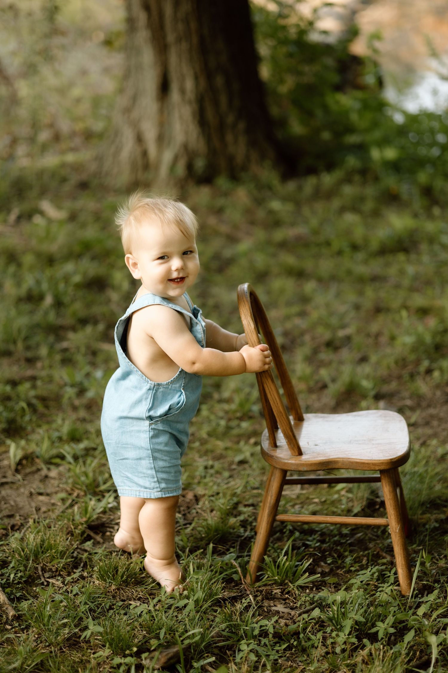 Smiling toddler in blue denim romper standing barefoot outdoors holding a wooden chair on green grass.