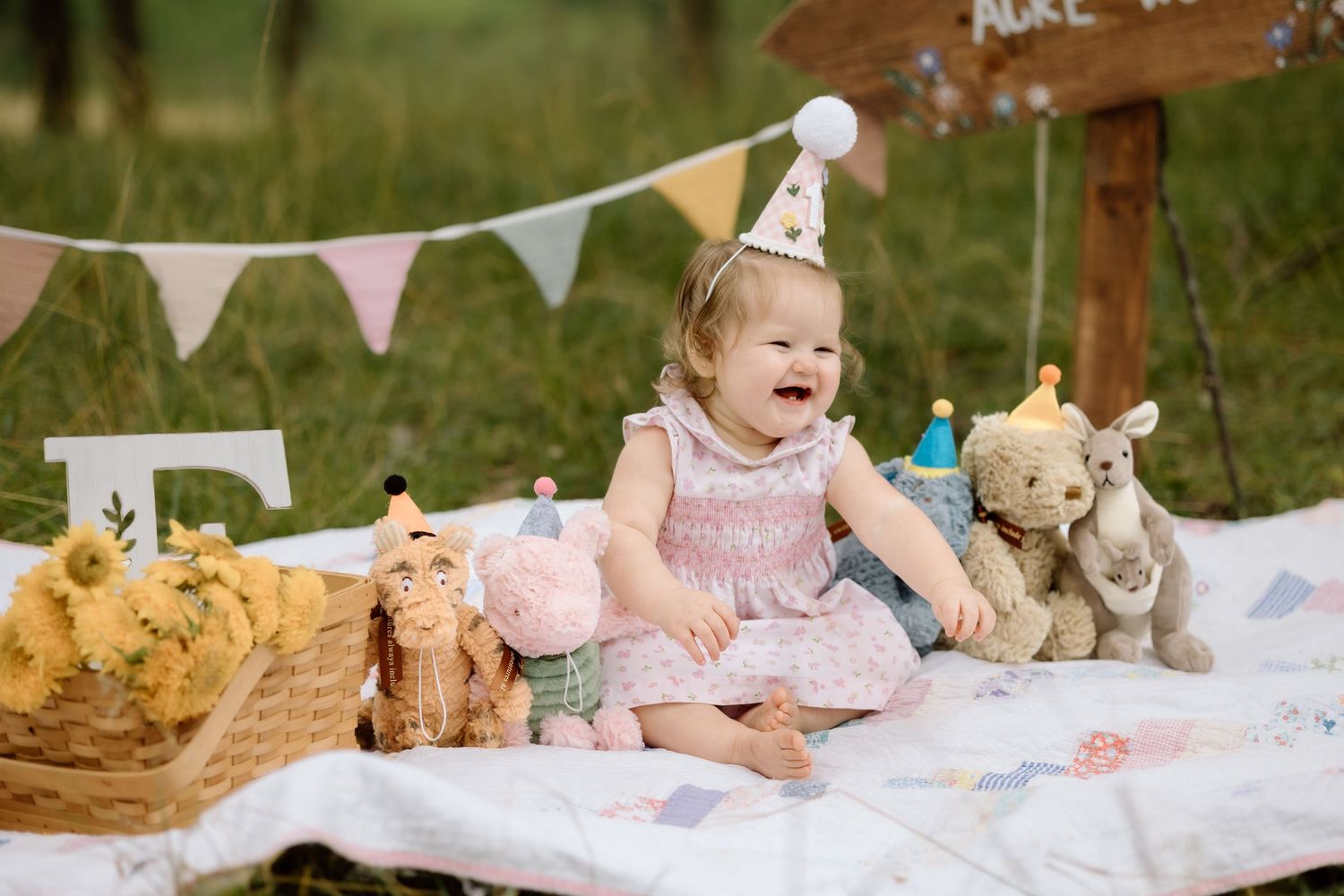 Happy baby girl in party hat laughs surrounded by stuffed animals at outdoor first birthday.