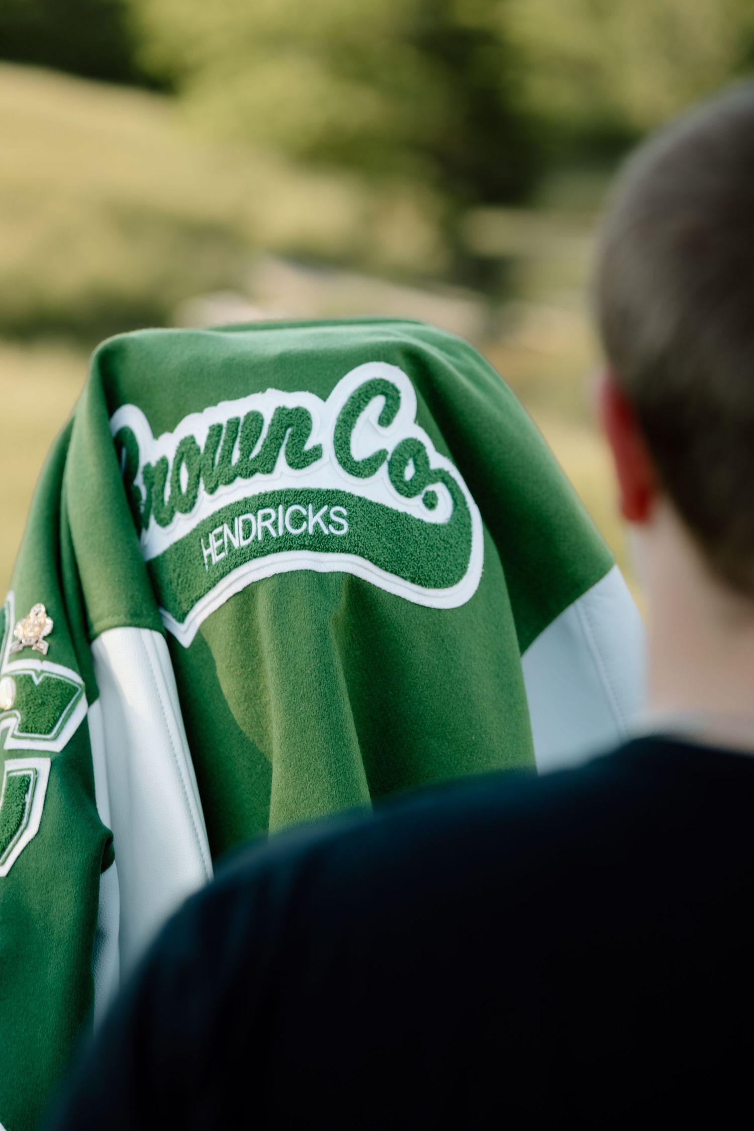 Back view of green Brown Co Hendricks letterman jacket draped over student's shoulder outdoors.