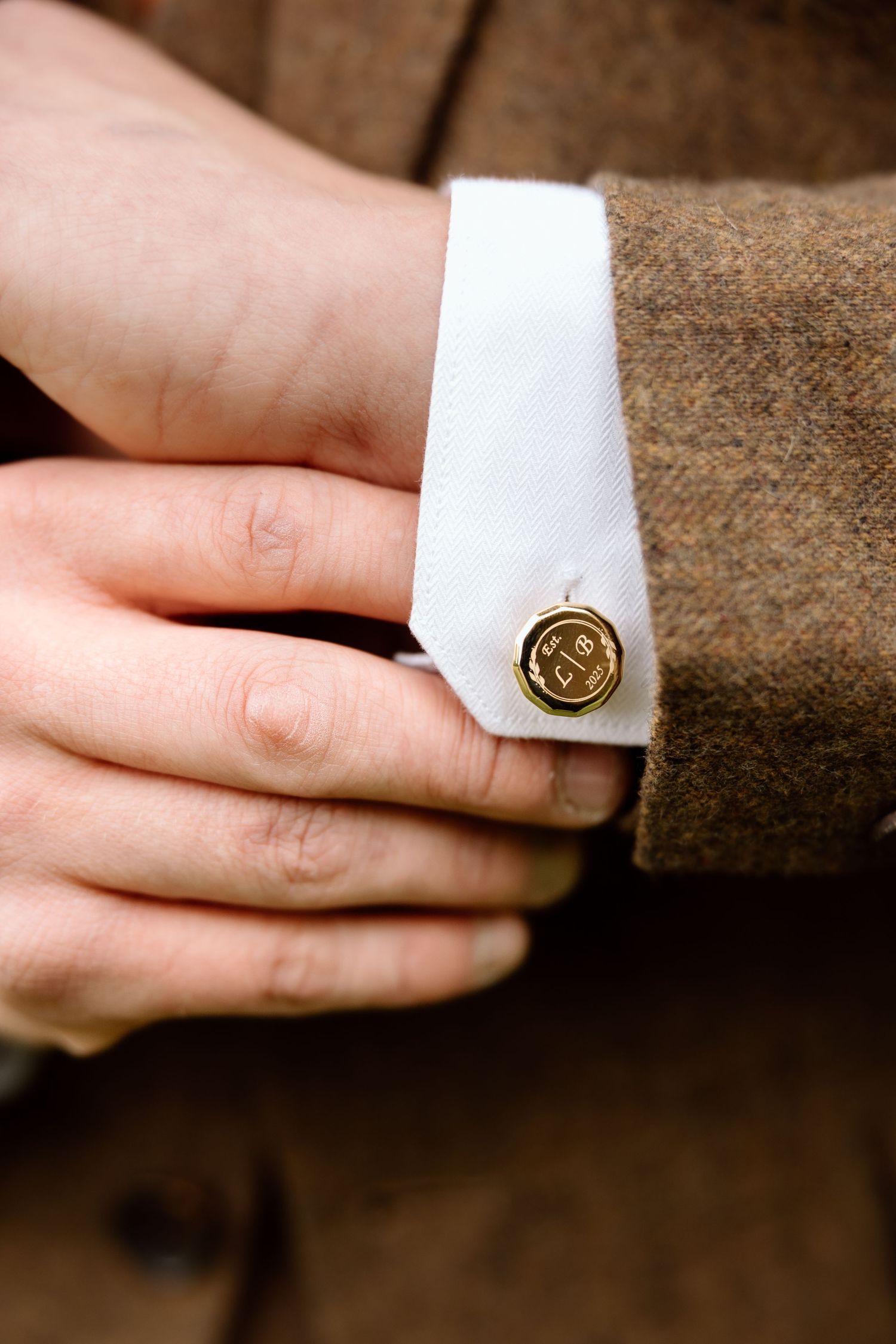 Close-up of gold monogrammed cufflink on white dress shirt cuff beneath brown tweed wedding suit jacket.
