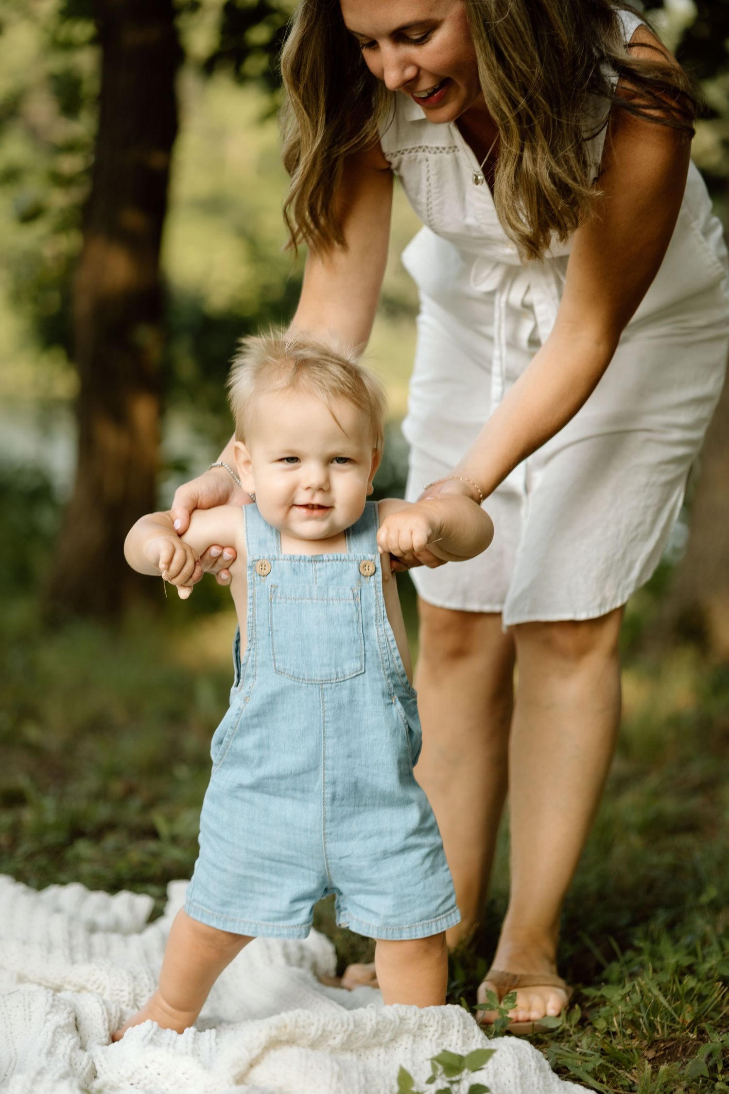 Mother in white dress helping baby boy take first steps on blanket outdoors.