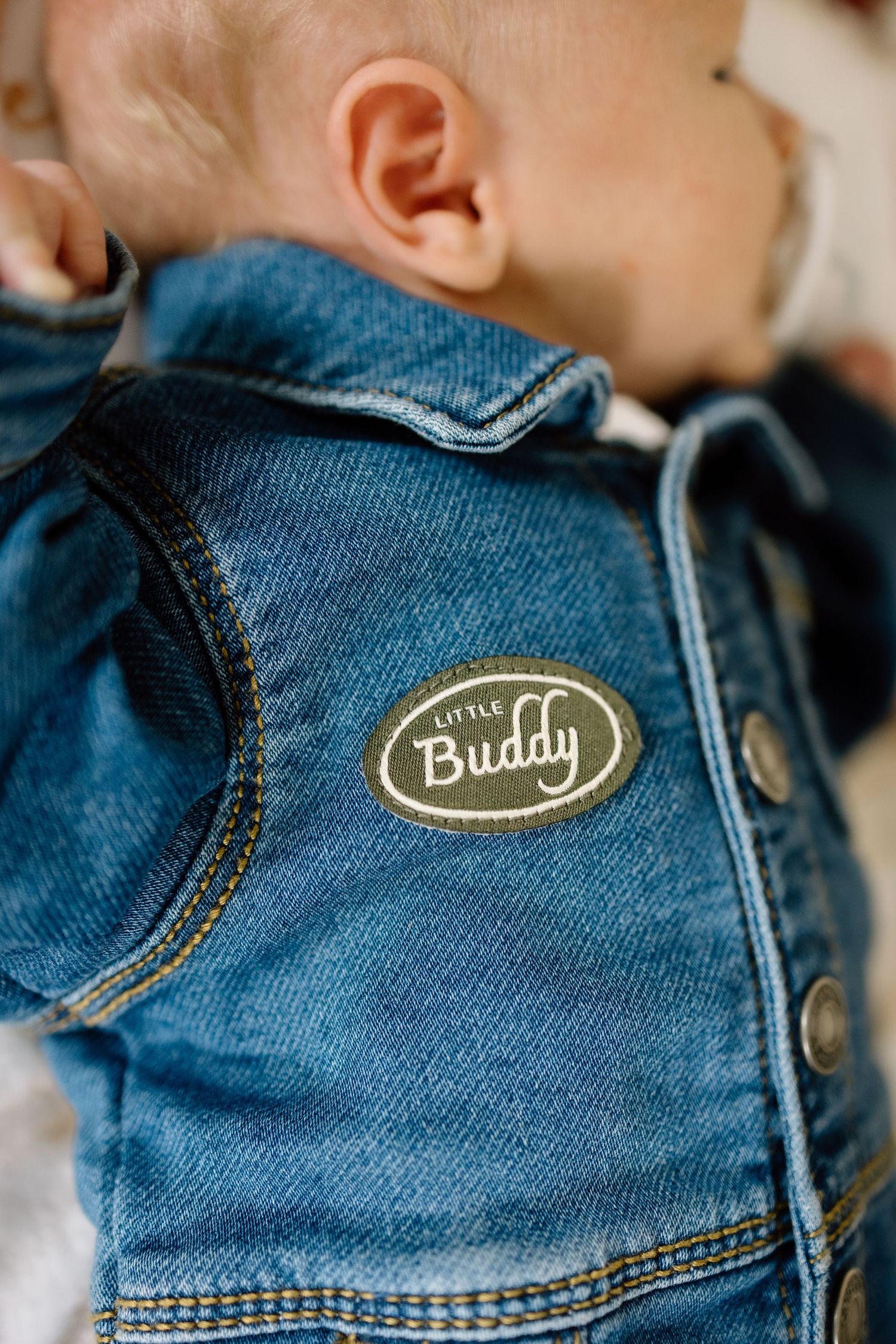 Close-up of a baby wearing a denim jacket with a Little Buddy patch on the shoulder.