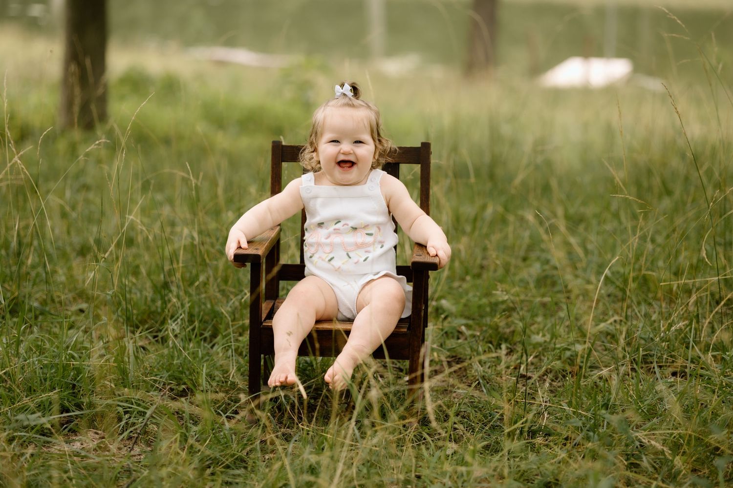 Laughing baby girl sits alone in a tiny wooden chair in a grassy outdoor field.