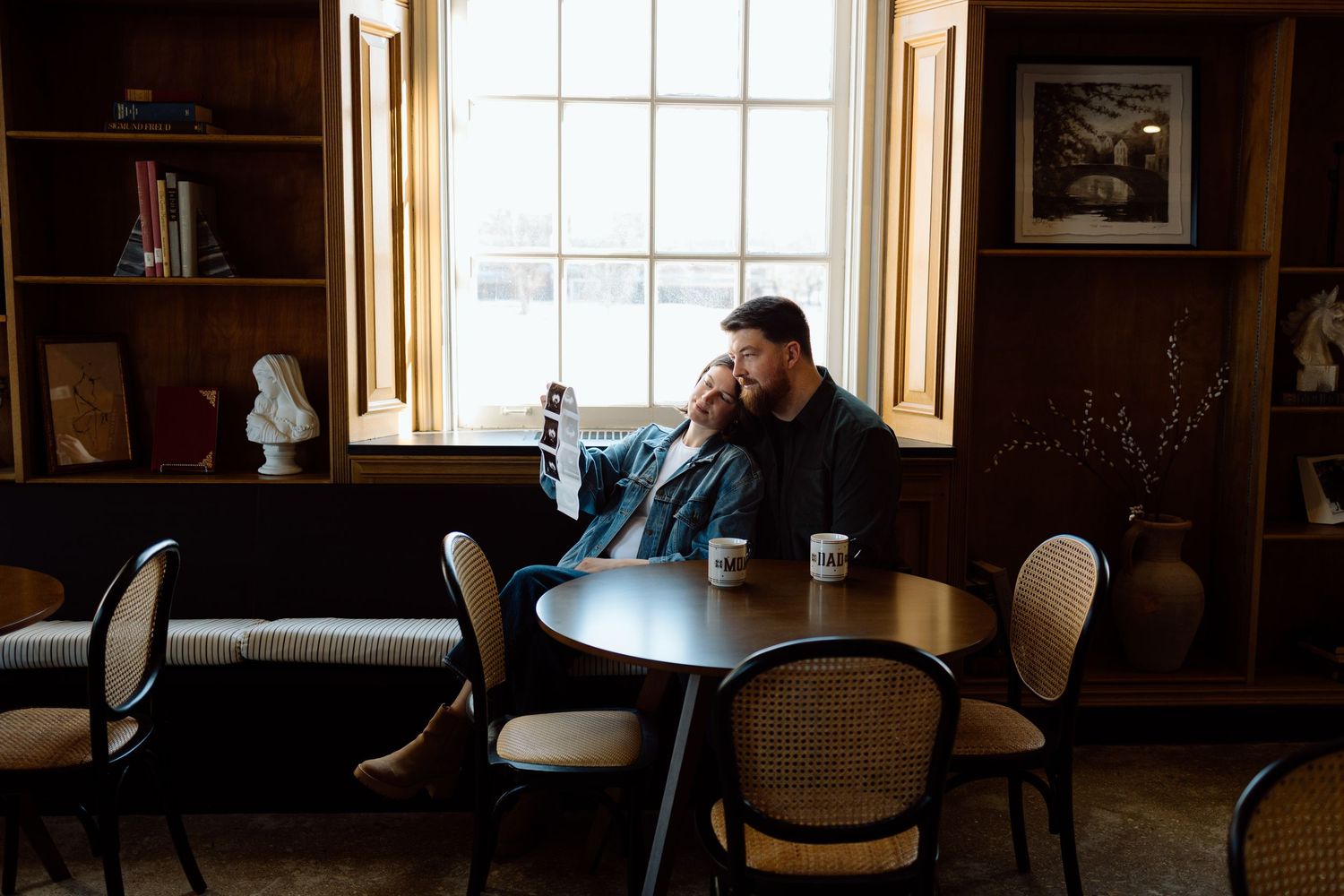 Bearded man sitting alone by window in cozy wood-paneled room holding coffee mug.