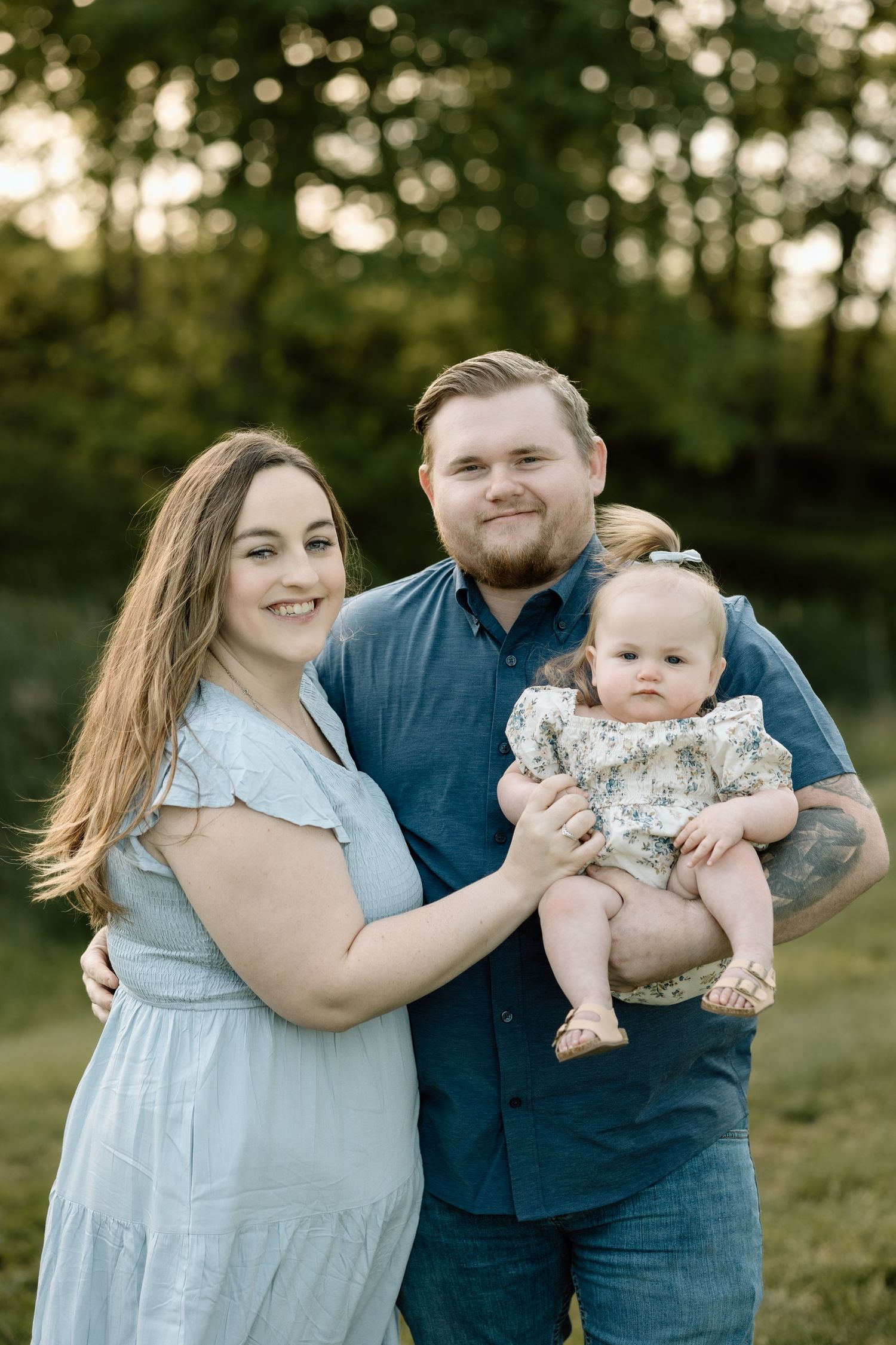 Smiling parents stand together in a sunny park, father holding their adorable baby girl in a floral outfit.