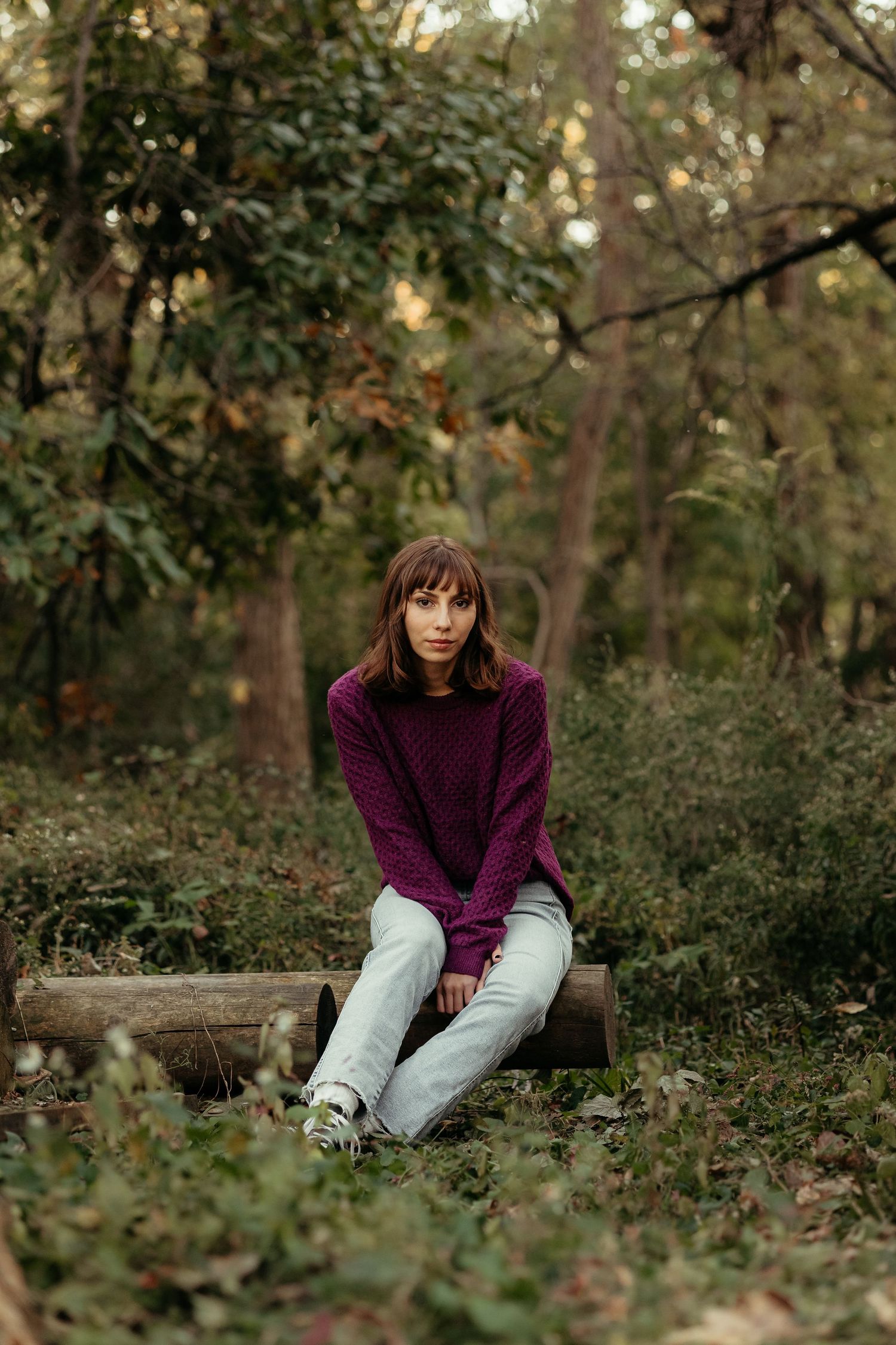 Young woman in purple sweater and jeans sits on a log in a lush green forest setting.