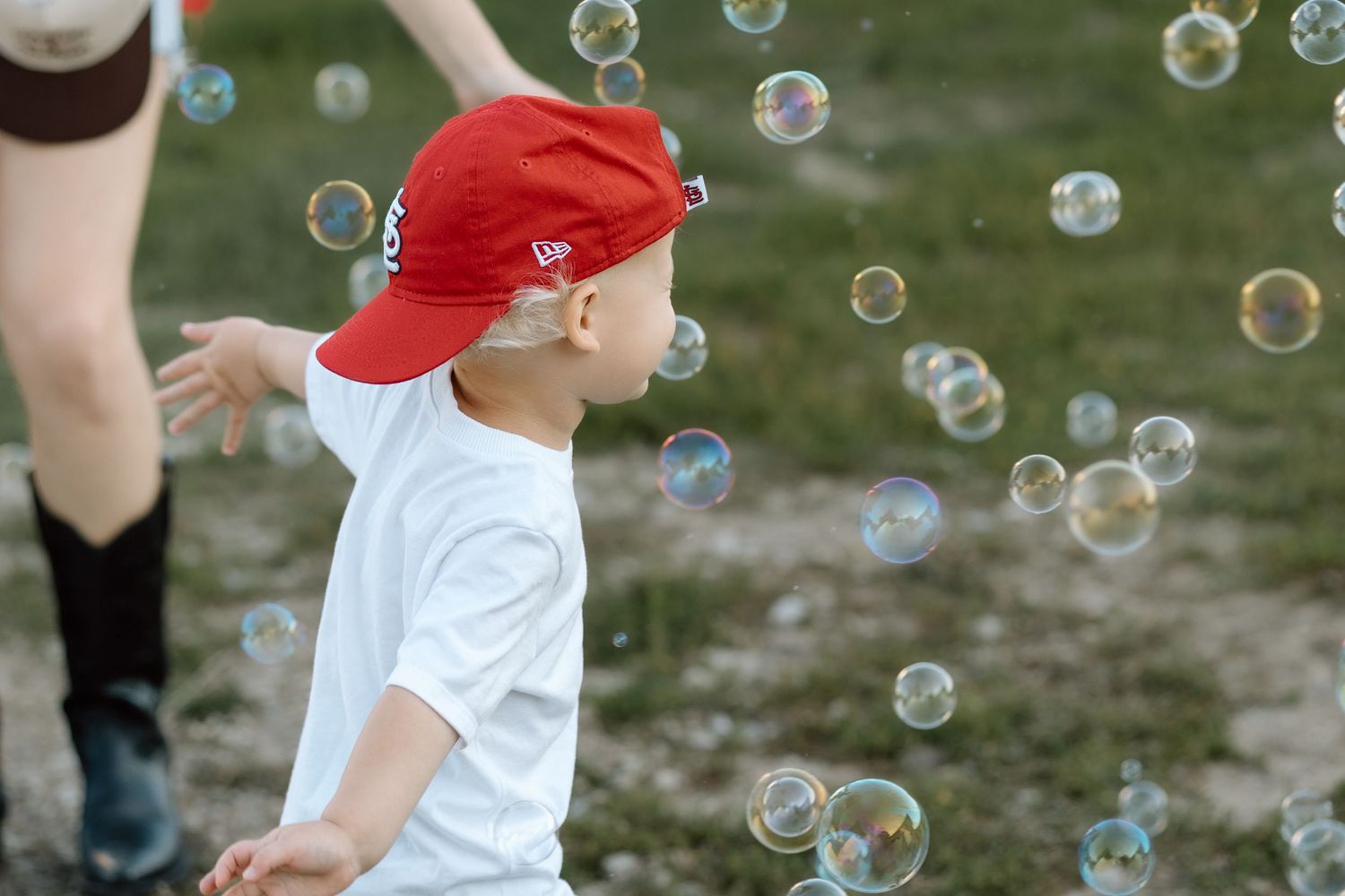 Toddler boy in red cap and white shirt playing with soap bubbles outdoors.