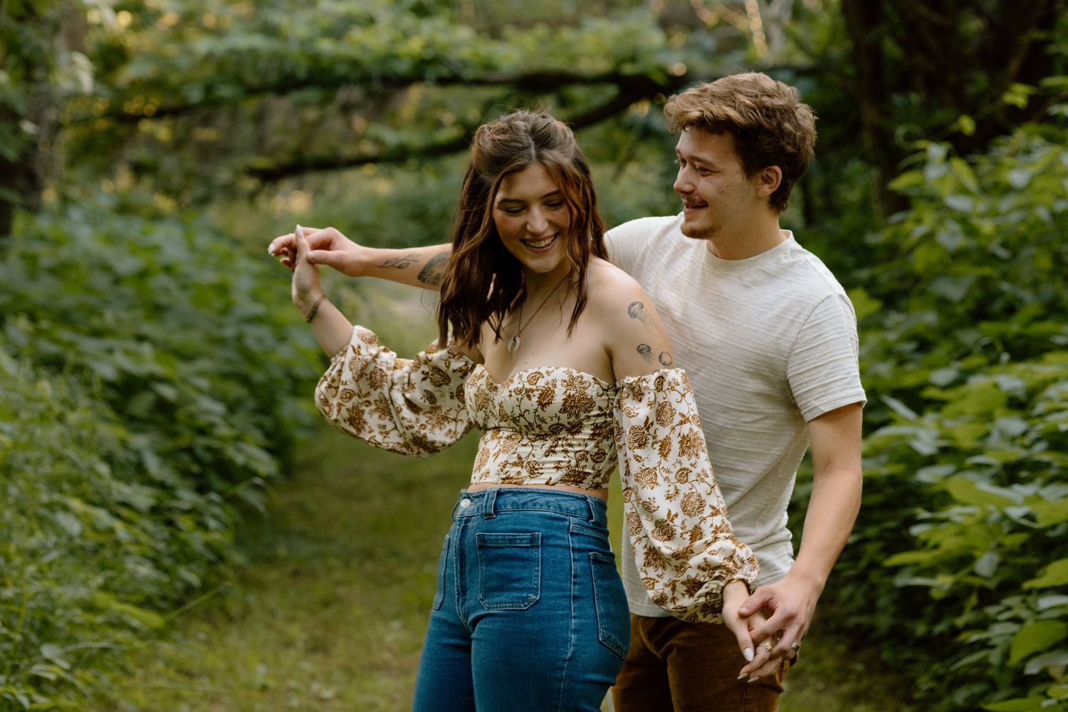 Happy couple holding hands and laughing while walking through a lush green wooded trail.