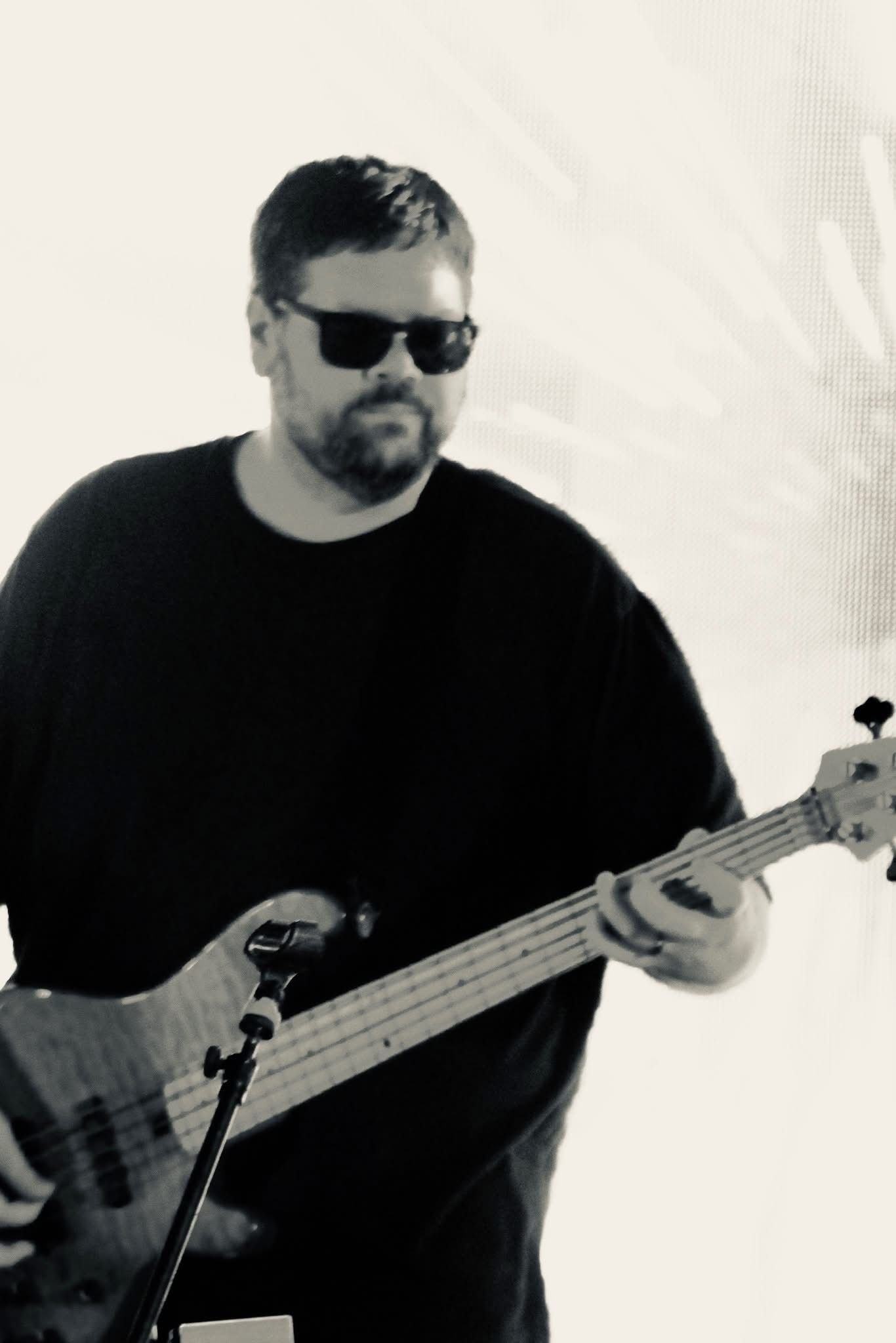 Black and white photo of a bearded man playing bass guitar wearing sunglasses outdoors.