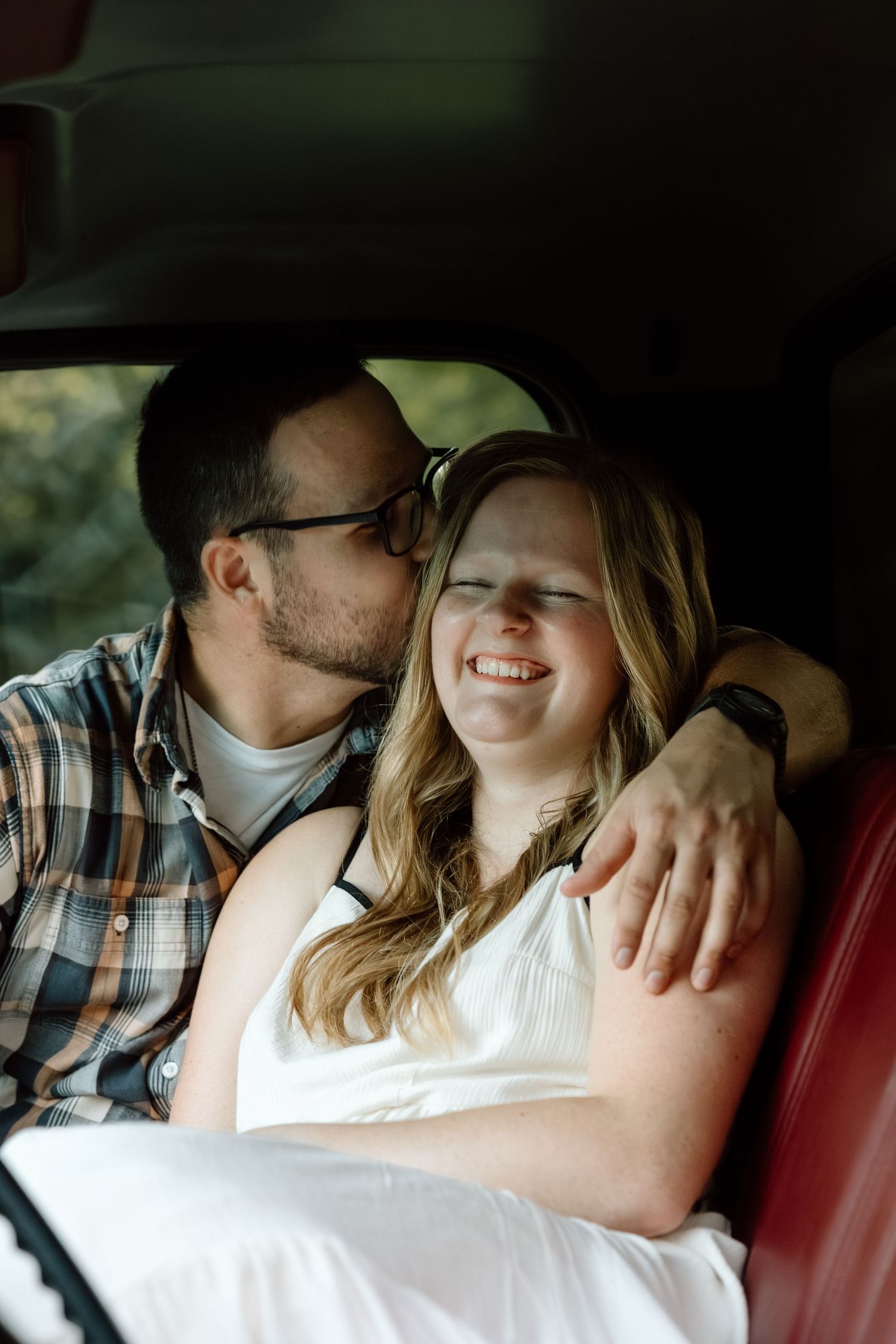 A couple shares a tender moment inside a vintage car with red leather seats, the man kissing the laughing woman on the cheek.