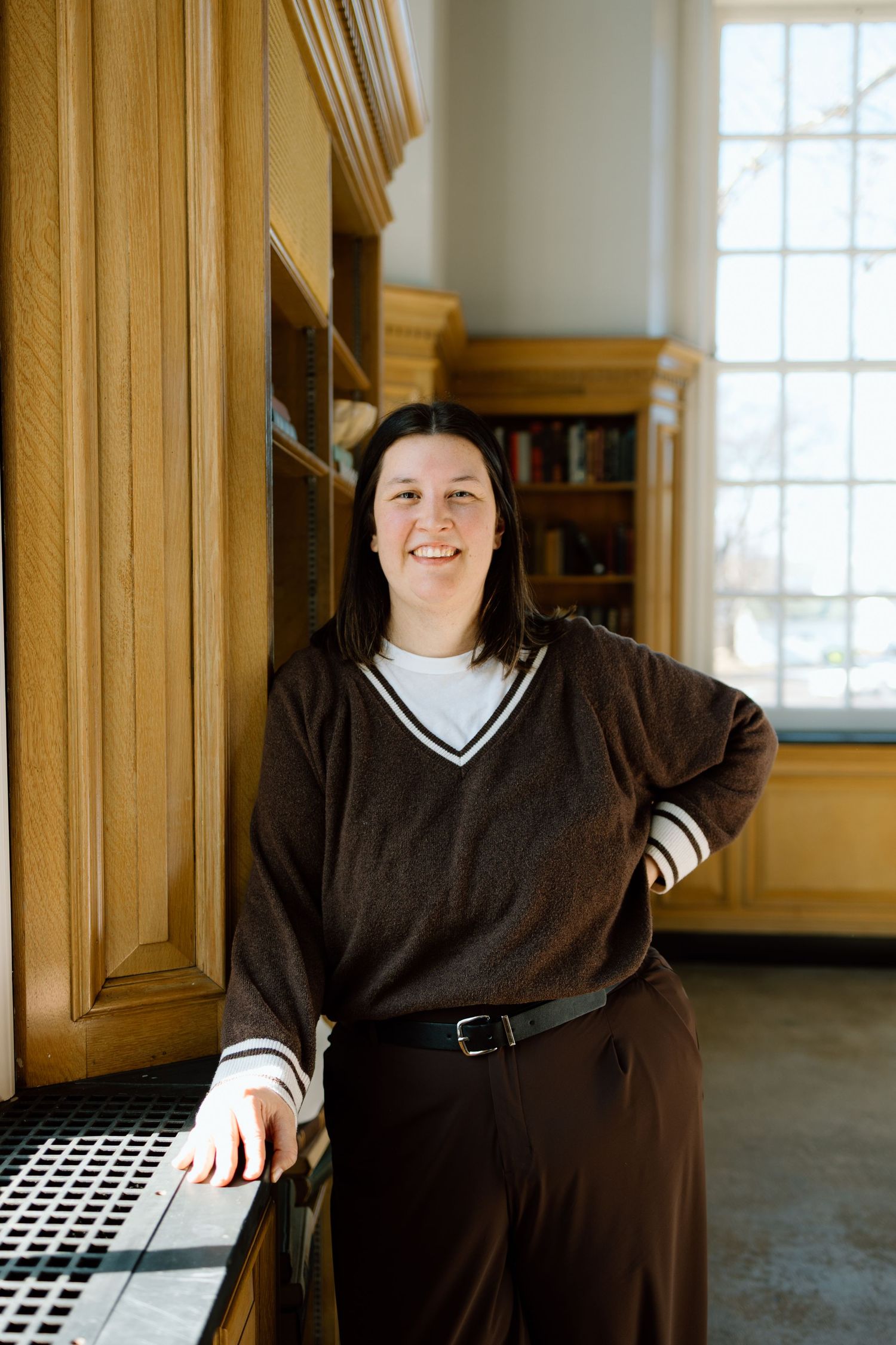 Smiling woman in brown V-neck sweater leaning against wooden cabinets in a bright library room.