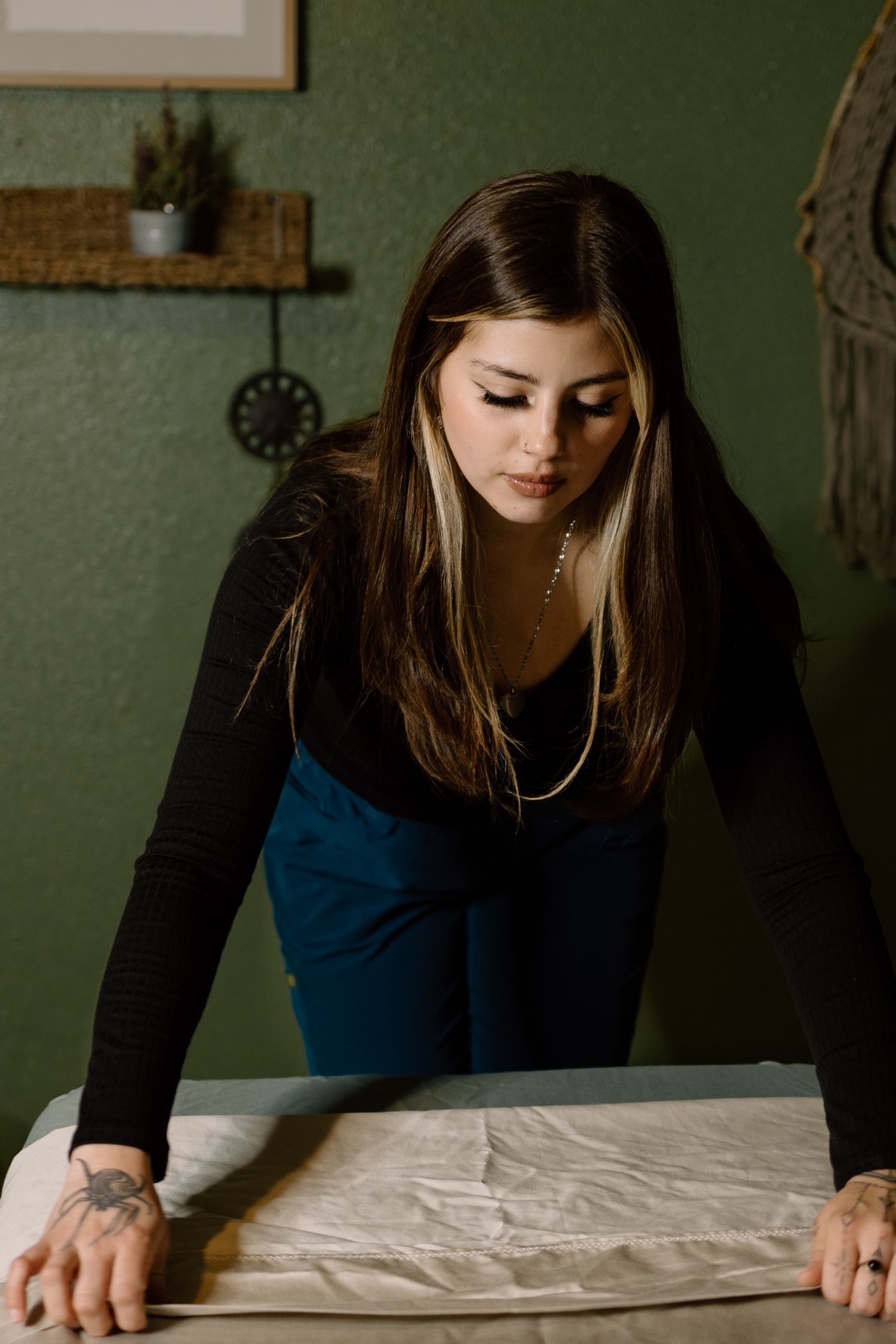 Young woman with highlighted hair in black top leaning over white massage table in green room.