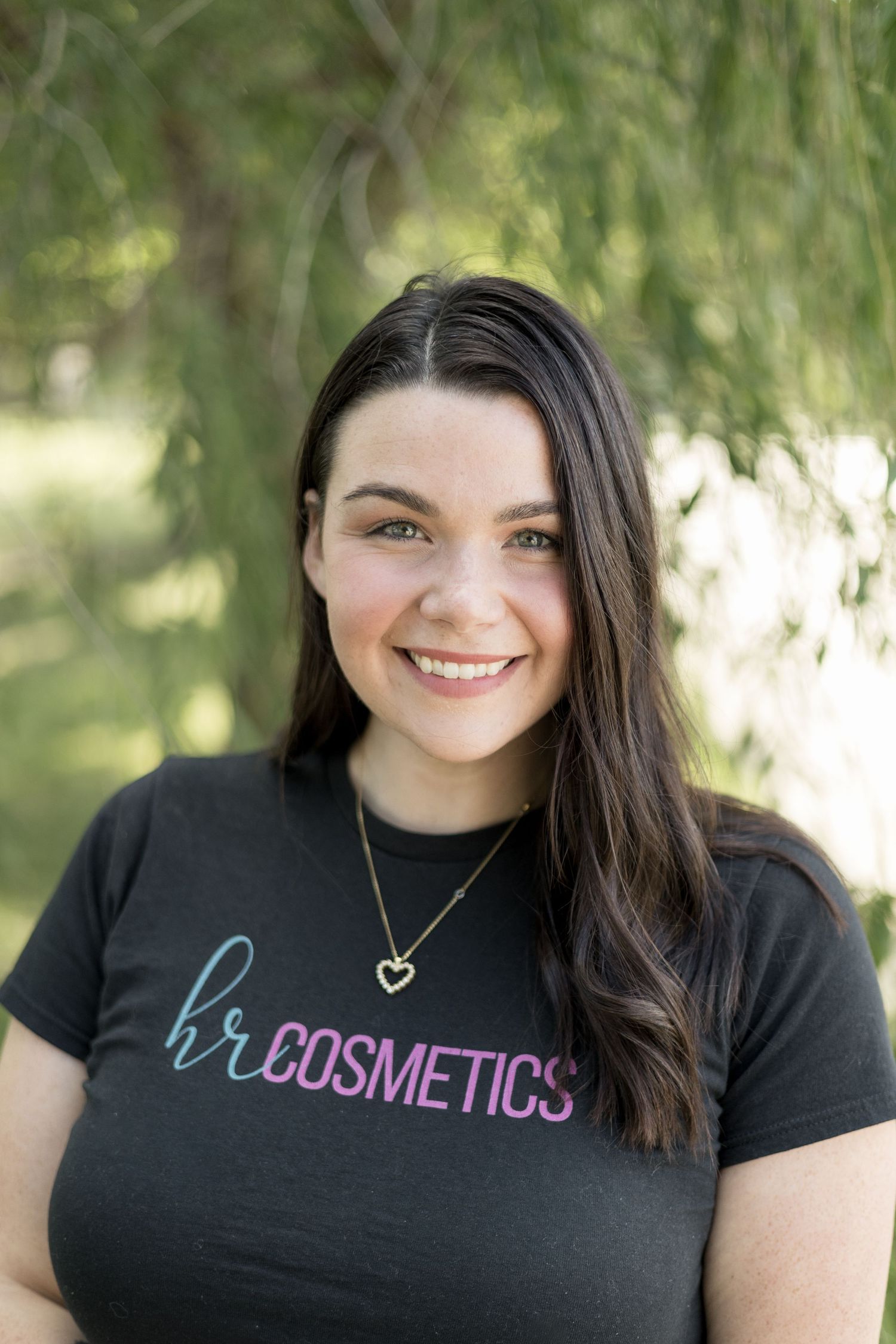 Headshot of smiling brunette woman wearing HR Cosmetics black t-shirt with gold heart necklace outdoors.