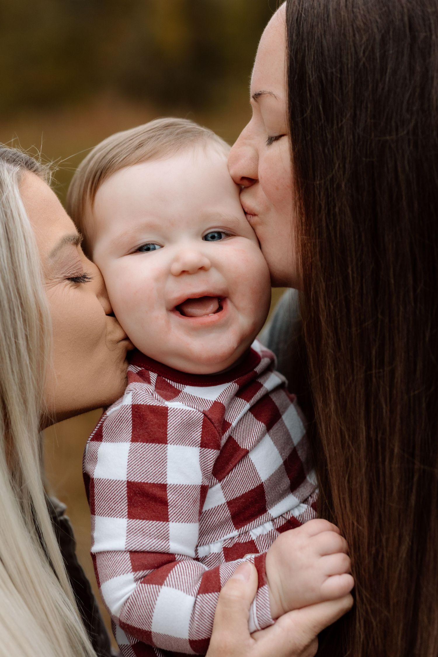 Two mothers kiss their giggling baby on the cheeks during an outdoor fall family portrait session.