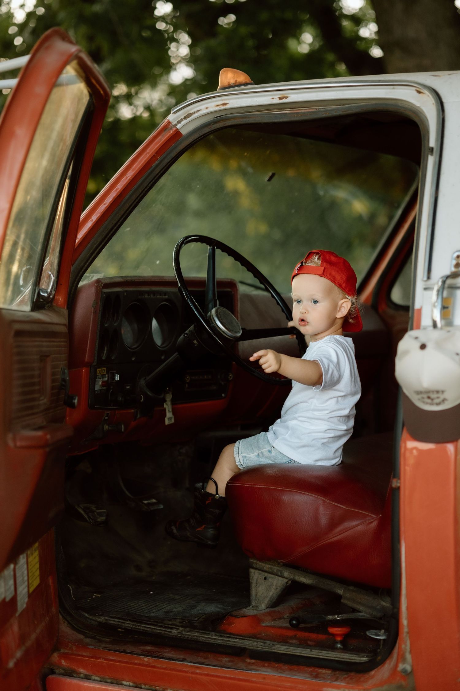 Cute toddler in red cap and white shirt sits in driver seat of old red farm truck.