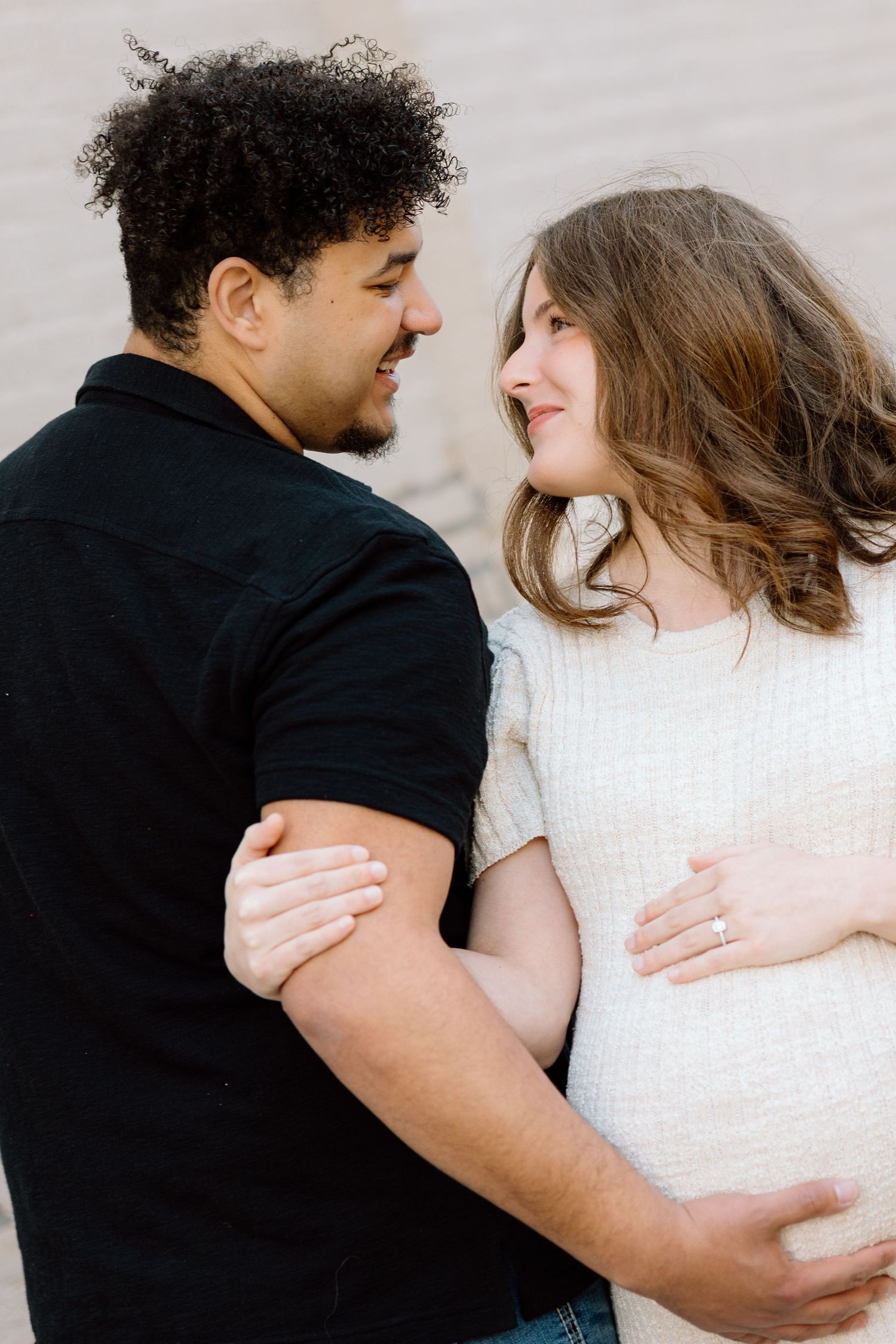 Happy interracial couple gazing at each other lovingly during outdoor maternity photoshoot near brick wall.