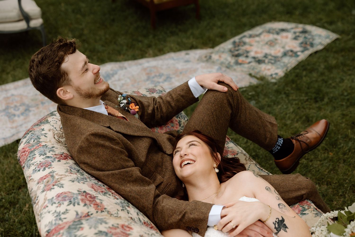 Laughing bride and groom lying together on a floral outdoor mattress during a relaxed wedding shoot.