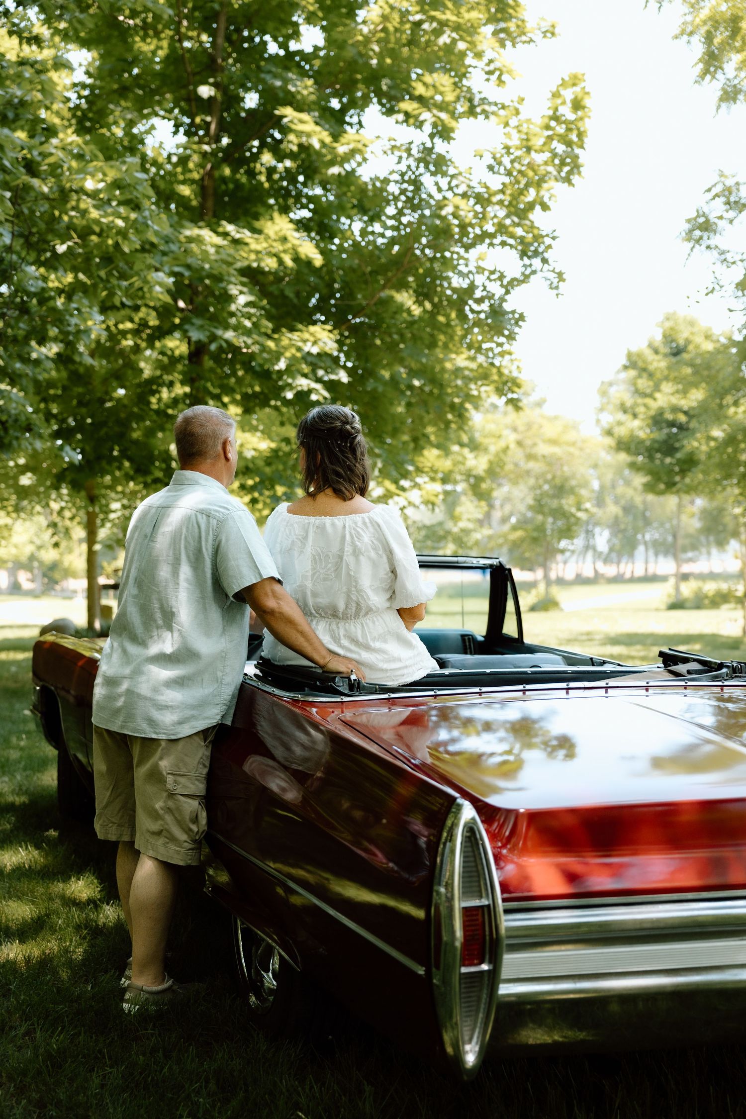 Bride and groom viewed from behind, standing beside a classic red convertible car in green park.