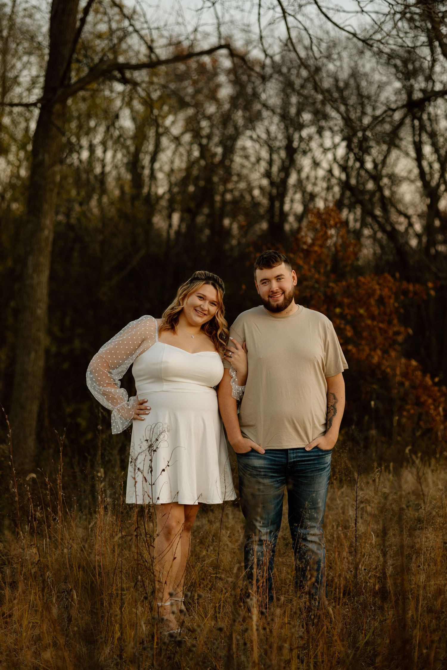 Couple posing together in autumn field at sunset, woman in white mini dress and man in tan shirt and jeans.