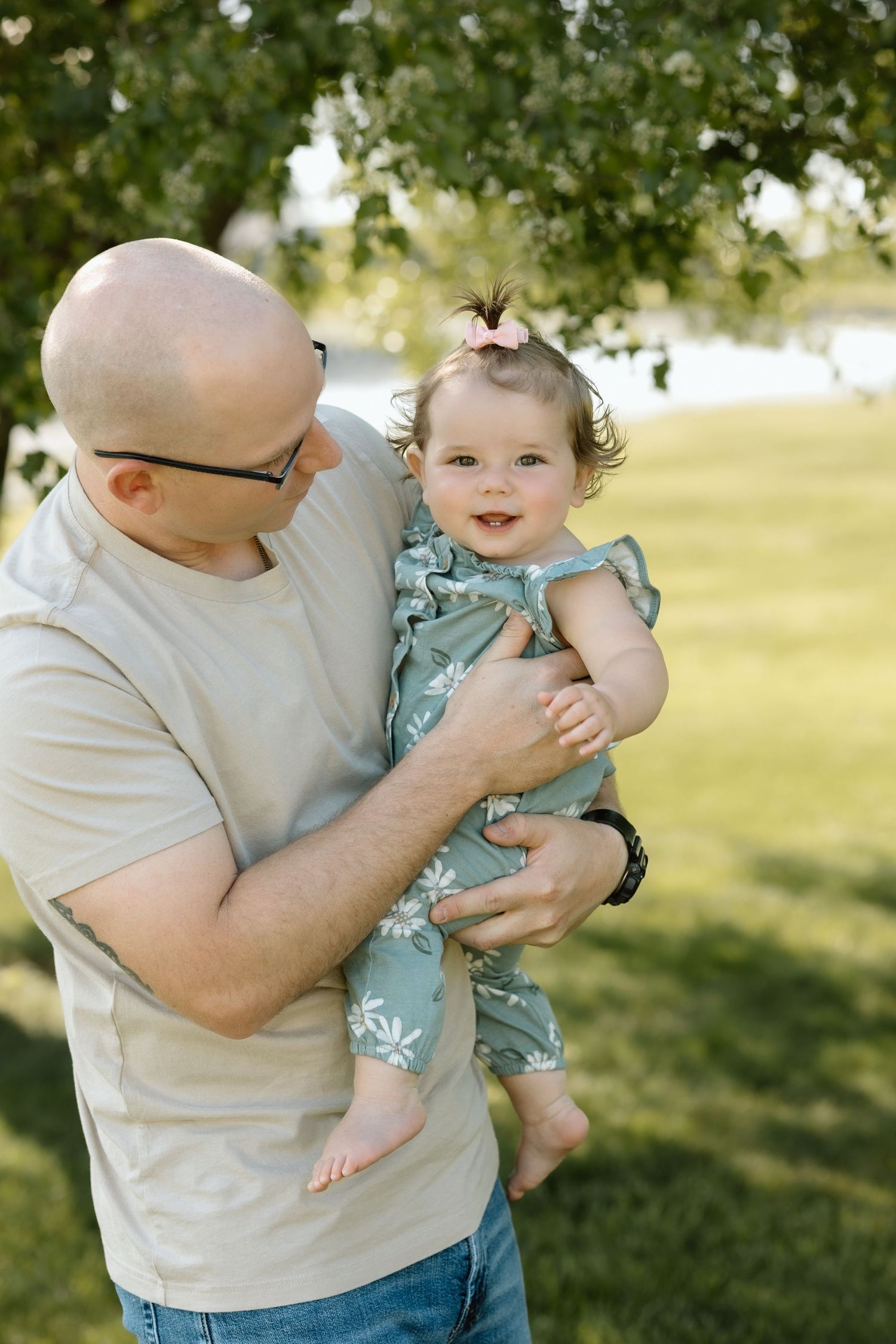 Bald father with glasses tenderly holds smiling baby girl in green floral romper outdoors in park.