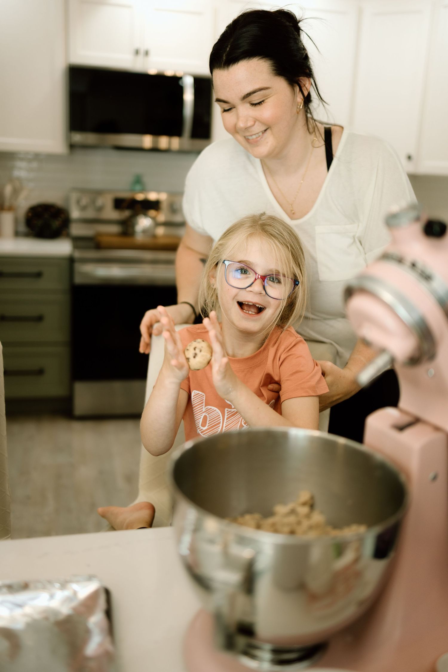 Excited young girl with glasses holding up cookie dough while mom smiles behind her at kitchen island.