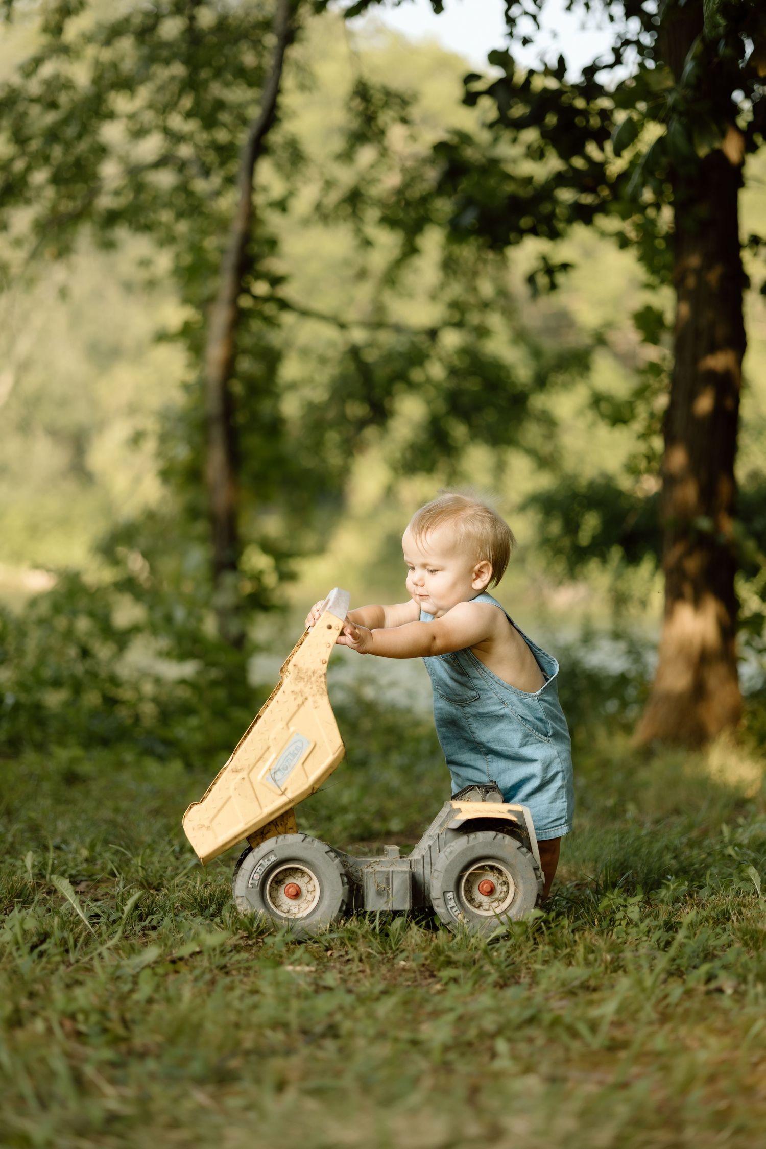 Toddler in blue denim romper playing with a yellow toy dump truck on green grass outdoors.