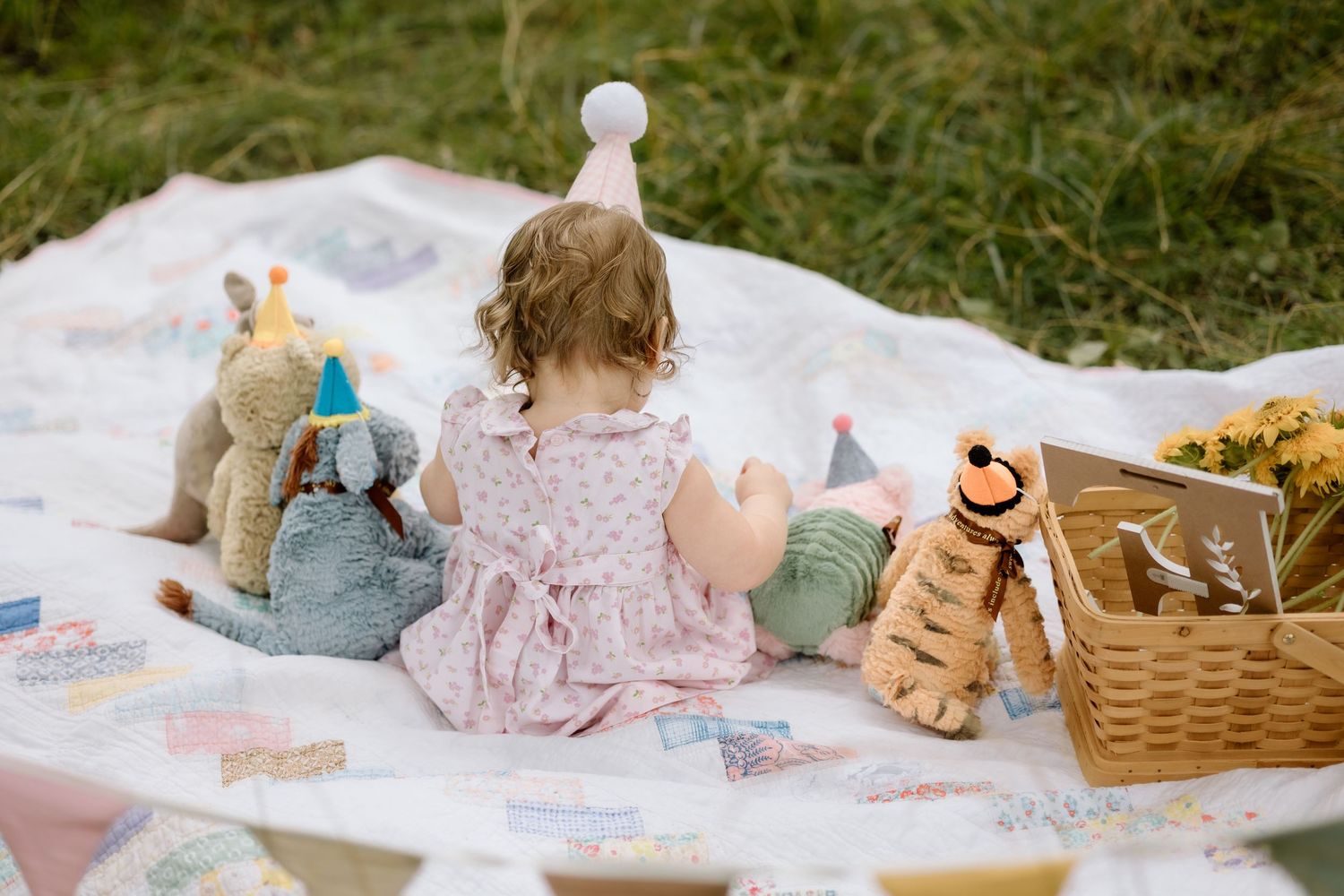 Baby girl in party hat sits with stuffed animals and a wicker basket on a picnic blanket.