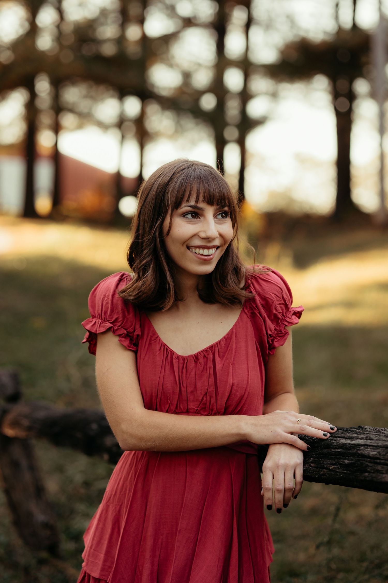 Smiling young woman in red dress leaning on a rustic wooden fence outdoors.