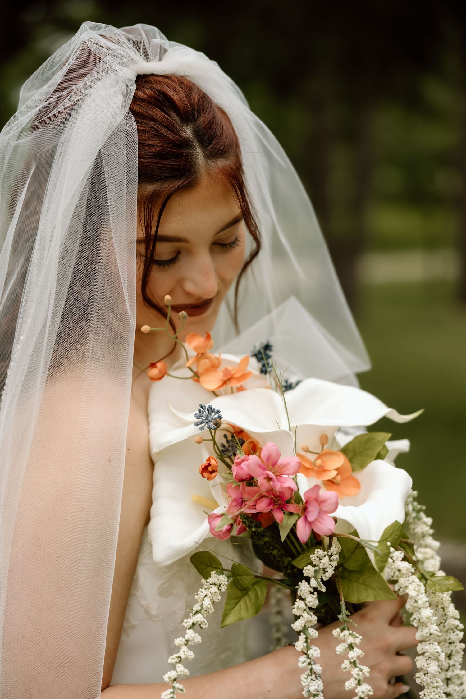 Bride with veil gazing down at colorful bouquet of calla lilies and orange and pink flowers.
