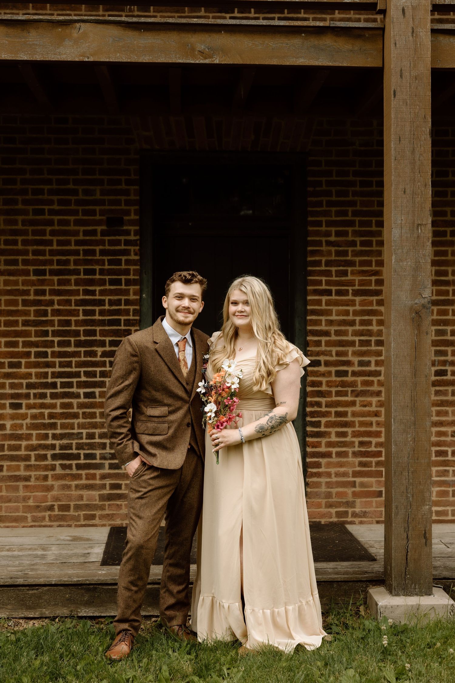 Wedding couple posing before brick building, groom in brown tweed suit, bride in champagne flowy dress.