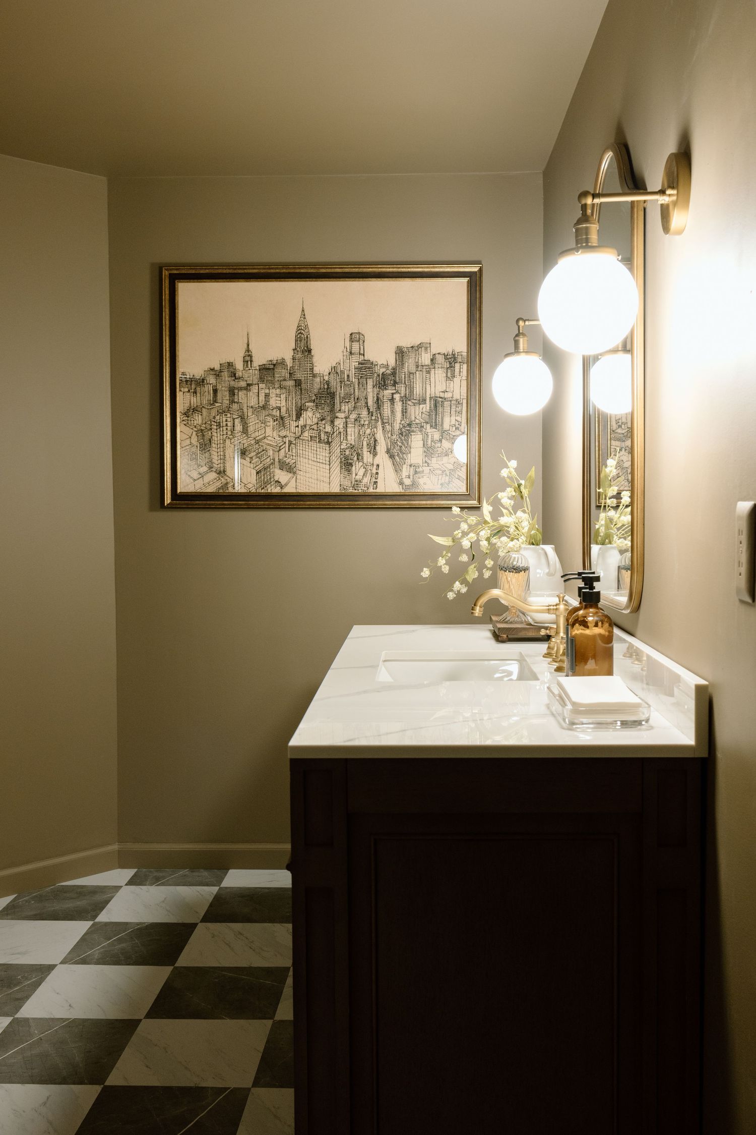Elegant bathroom vanity with dark wood cabinets, marble countertop, gold fixtures, checkered tile floor, and NYC skyline artwork.