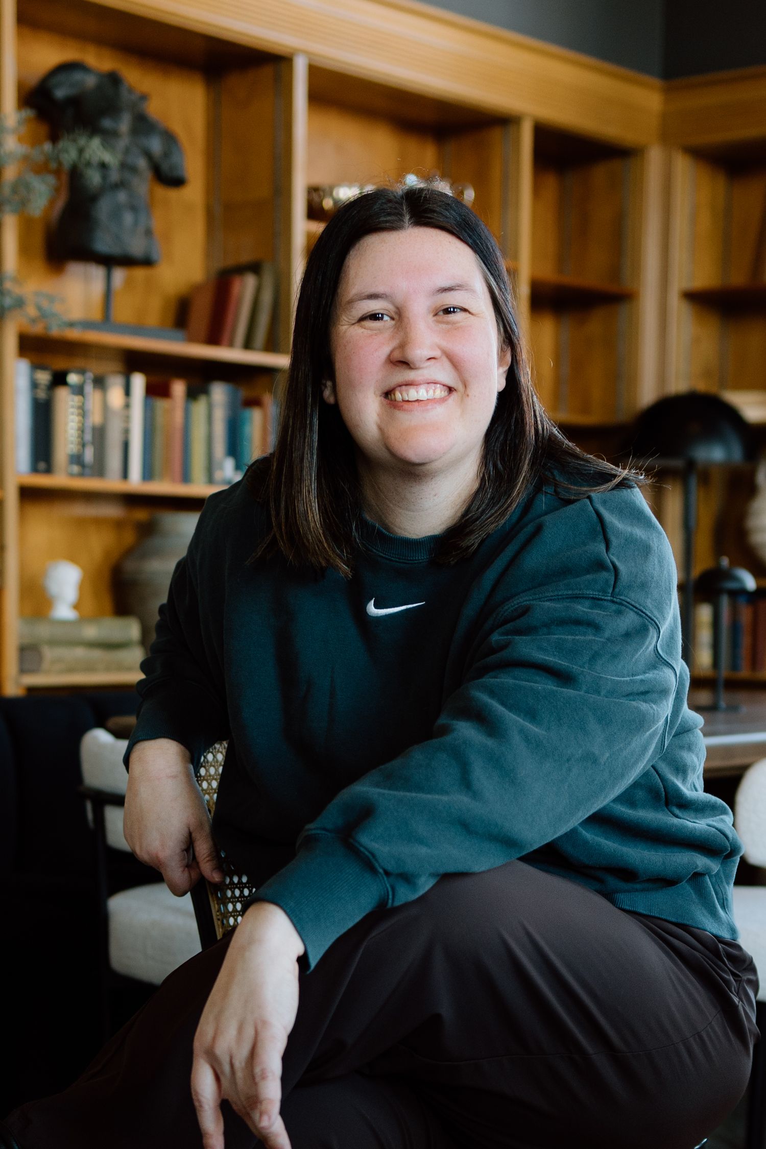 Smiling woman in teal Nike sweatshirt sitting in front of wooden bookshelf filled with books.