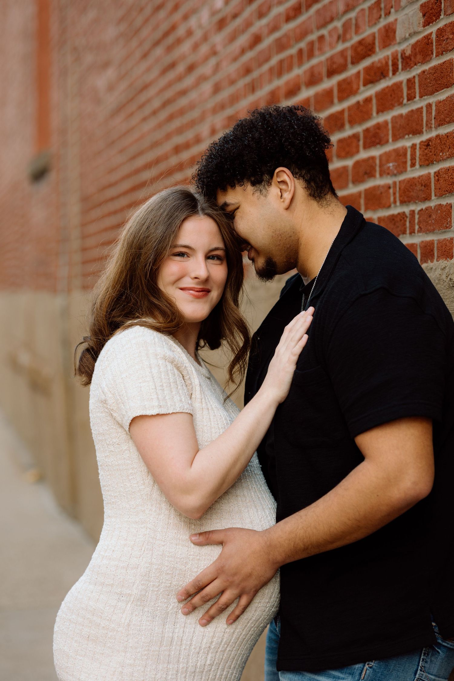 Expecting couple sharing a tender moment against a red brick wall, man gently touching pregnant belly.