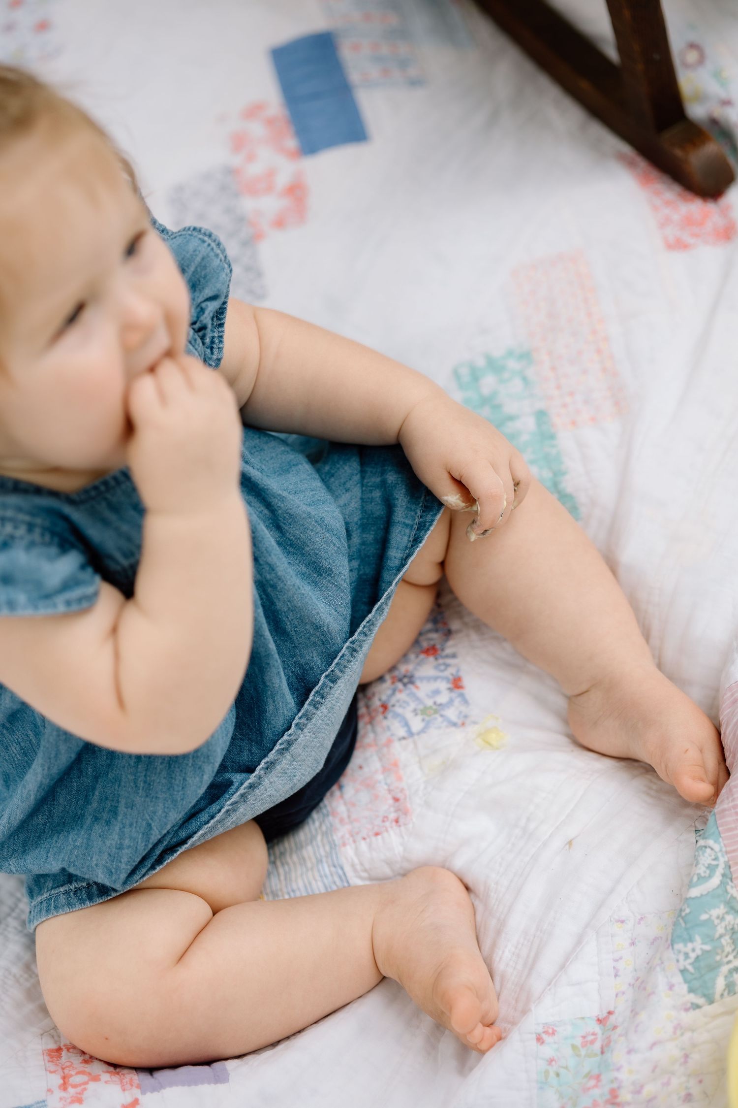 Cute baby girl in denim dress sitting on colorful patchwork quilt with finger in mouth.