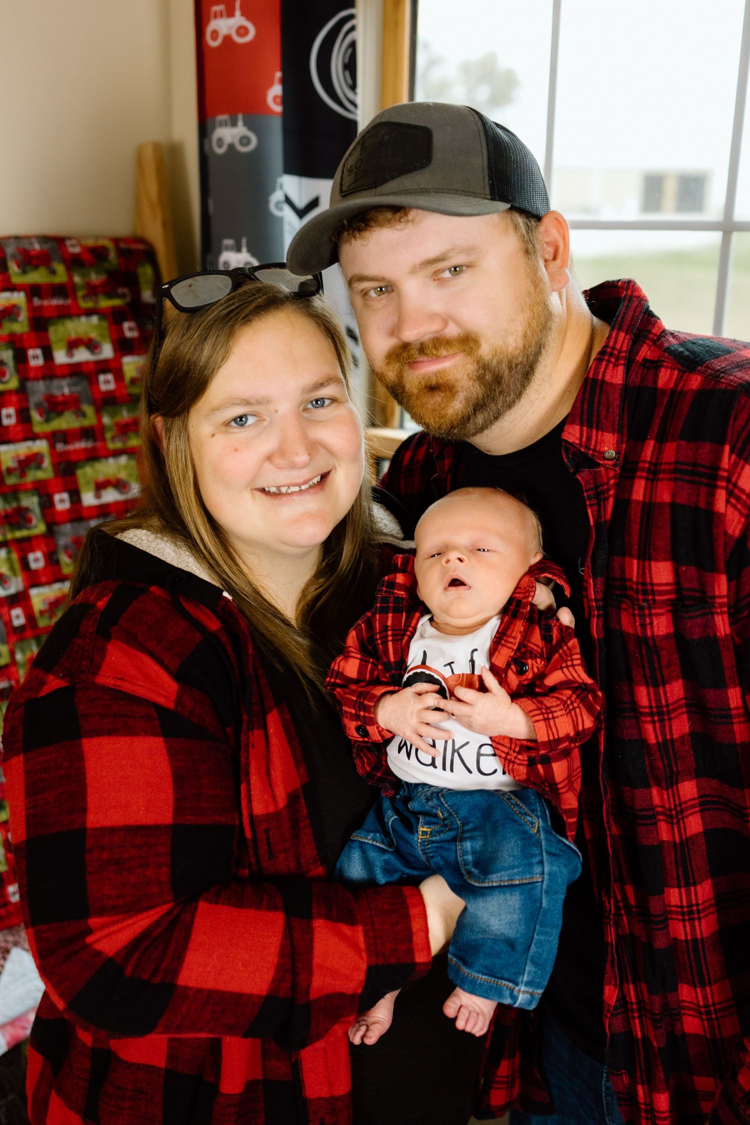 Smiling family in matching red plaid shirts posing together with their newborn baby.