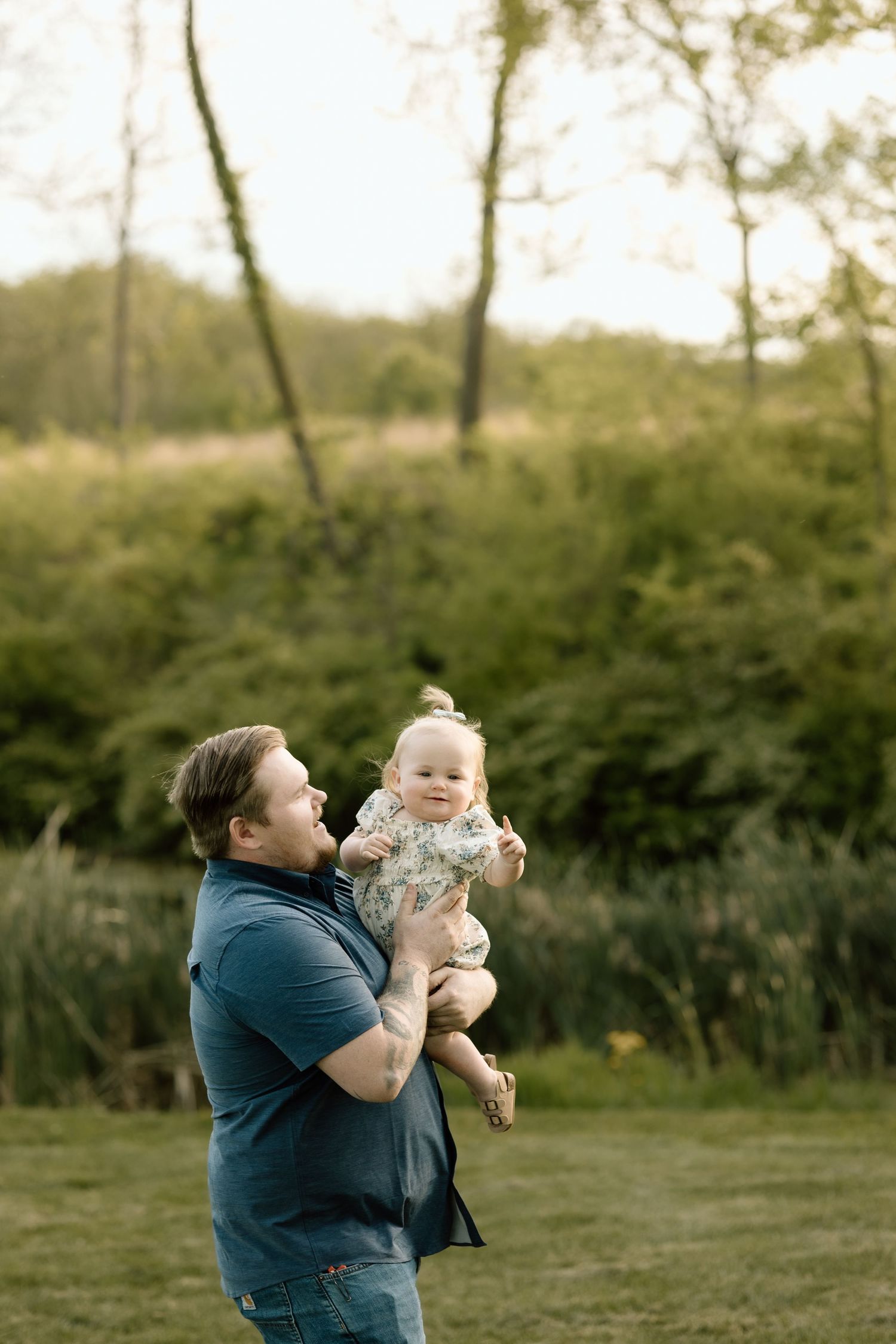 A proud father lifts his happy baby girl up in the air outdoors in a lush green park at golden hour.