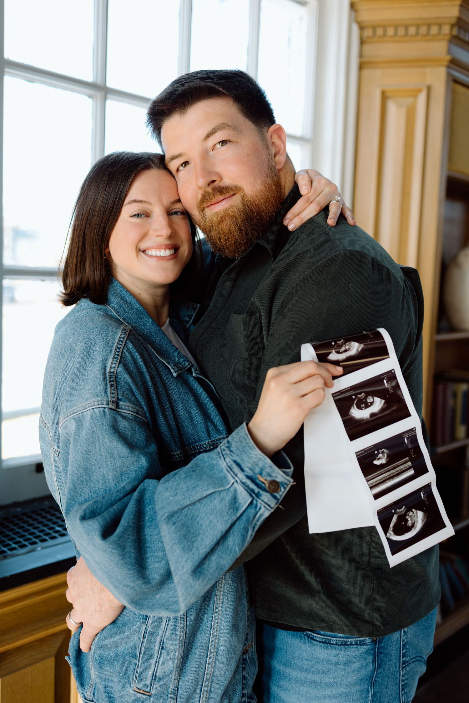 Smiling couple poses together holding ultrasound images during their pregnancy announcement photoshoot.