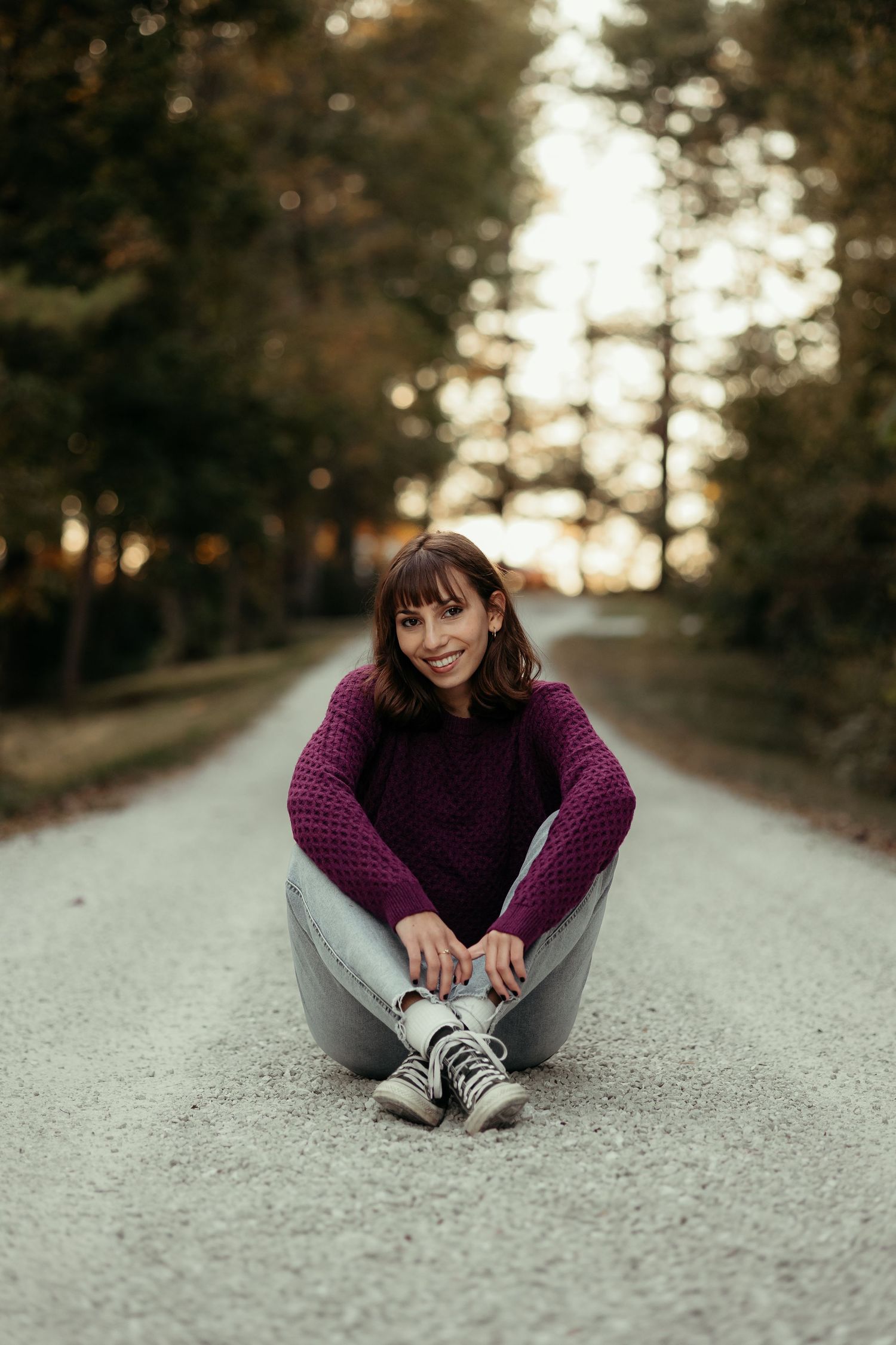 Smiling young woman in purple sweater sits cross-legged on a gravel road in autumn forest.
