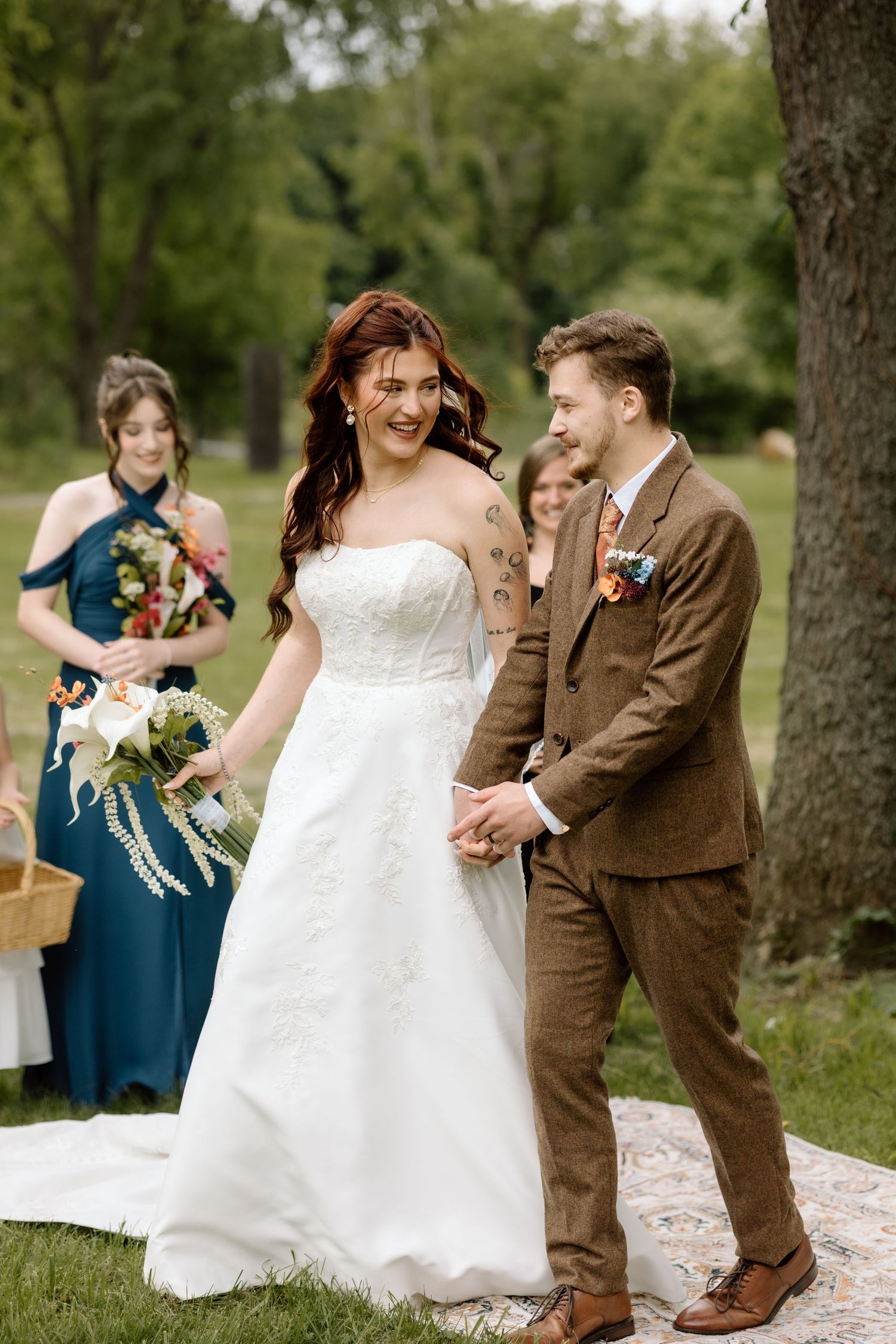 Bride in white strapless gown and groom in brown tweed suit hold hands smiling during outdoor ceremony.