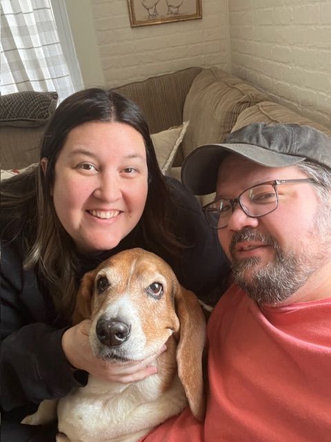 A smiling couple poses on a couch with their happy basset hound dog looking directly at the camera.