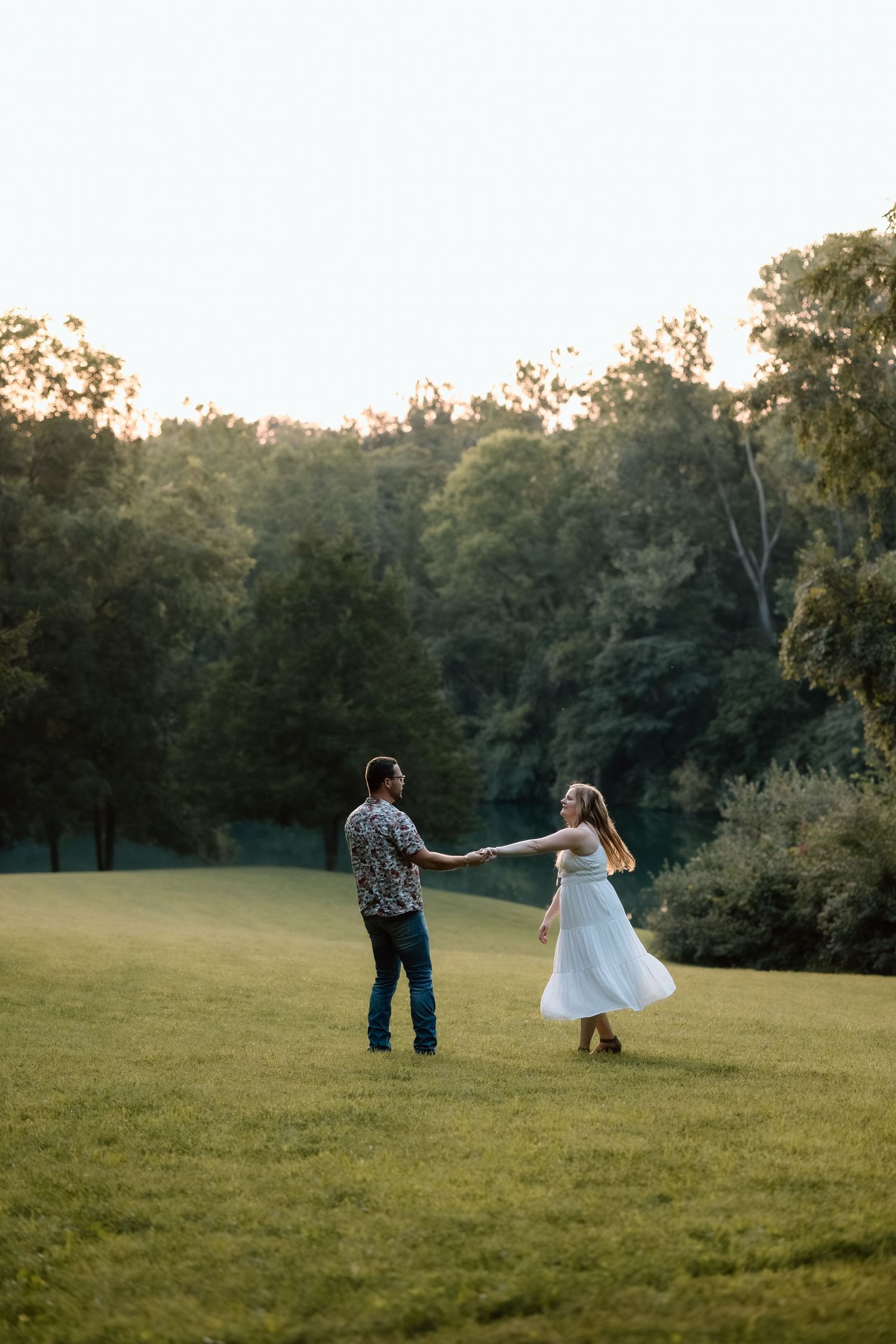 A couple dances and twirls on a lush green lawn at sunset, surrounded by tall trees in warm golden light.