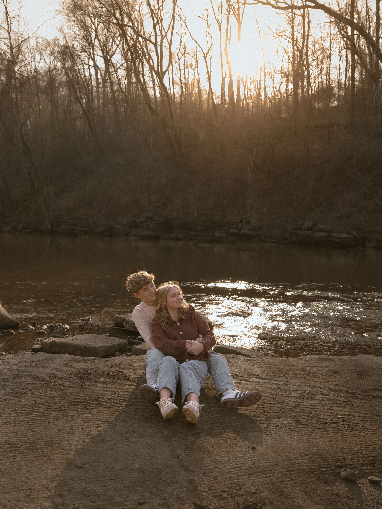 Couple sitting together on rocks by a sunlit river at sunset, sharing a tender moment in a wooded setting.