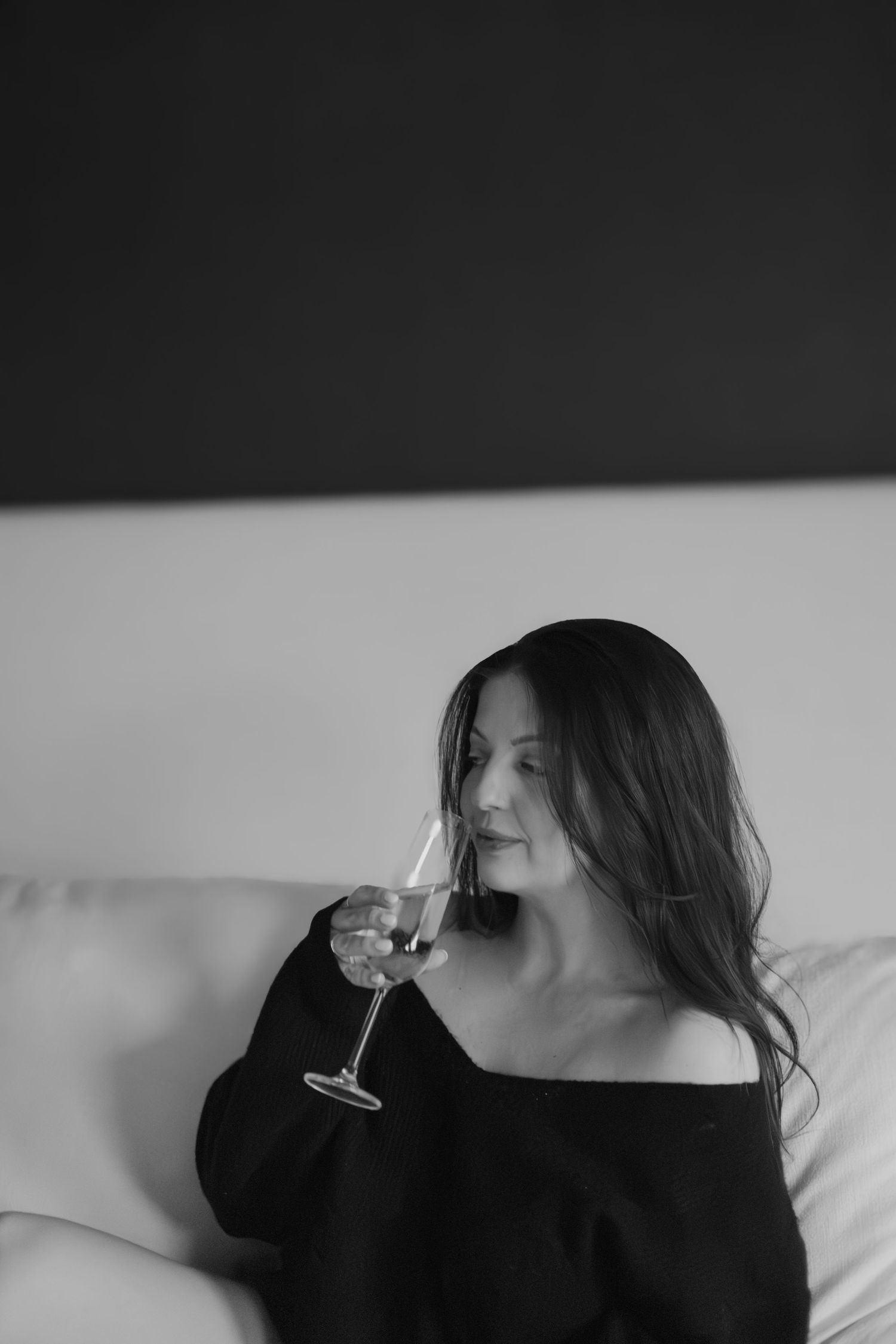 Black and white photo of a woman with long dark hair holding a wine glass while relaxing on a white sofa.